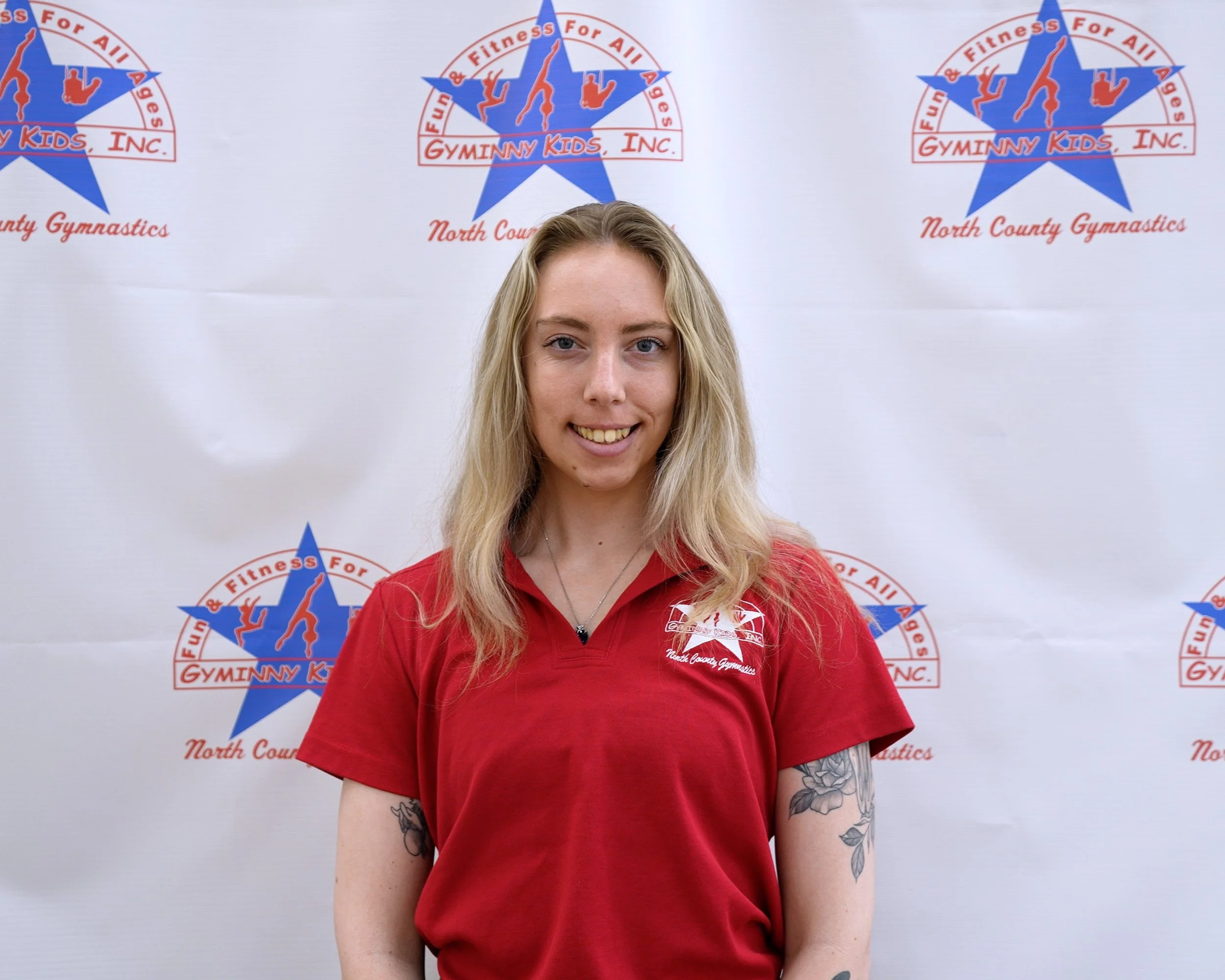 A young woman with long blonde hair wearing a red shirt with a gymnastics logo in front of a backdrop with gymnastic-themed logos and text for North County Gymnastics.