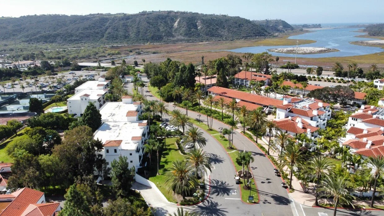 Aerial view of a coastal residential area with white apartment buildings and red-tiled roofs, lined with palm trees, near a body of water and a marshland with hills in the background.