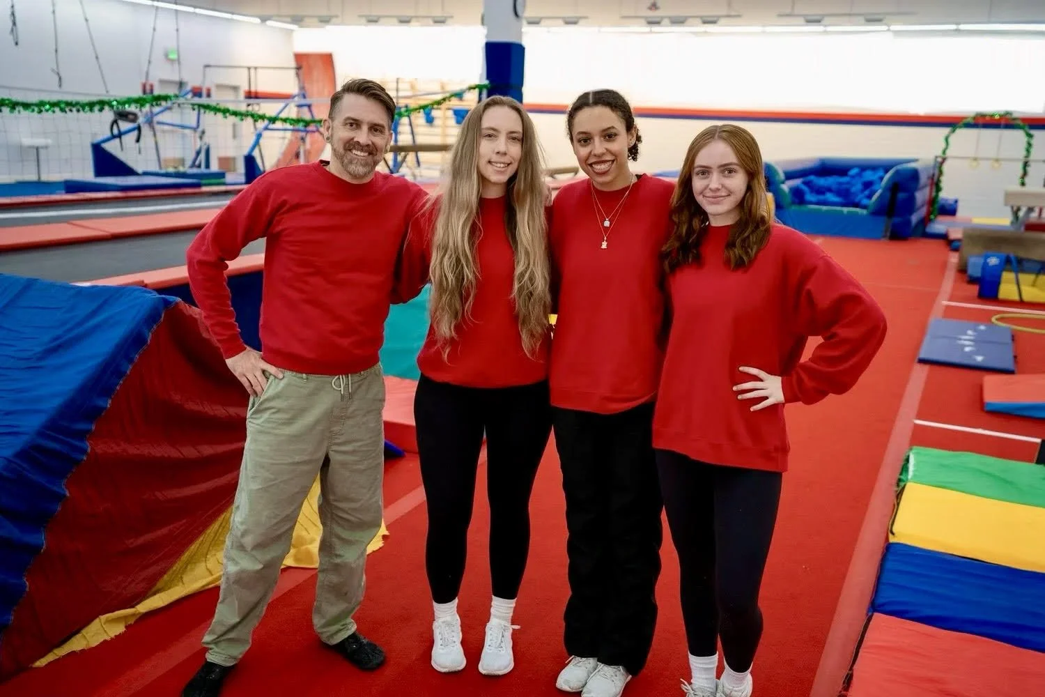 Four people standing in an indoor trampoline park, all wearing red shirts and black or khaki pants, smiling at the camera. The background shows colorful trampolines and a sign that reads 'Spring Break & Summer Camp'.