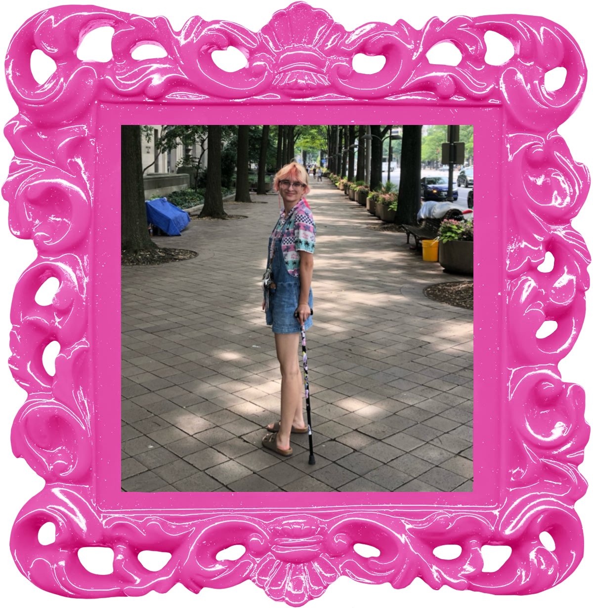 A person with light pink hair and glasses, wearing a colorful patterned shirt and denim shorts, stands on a shaded city sidewalk using a cane. The photo is in a curling pink frame.