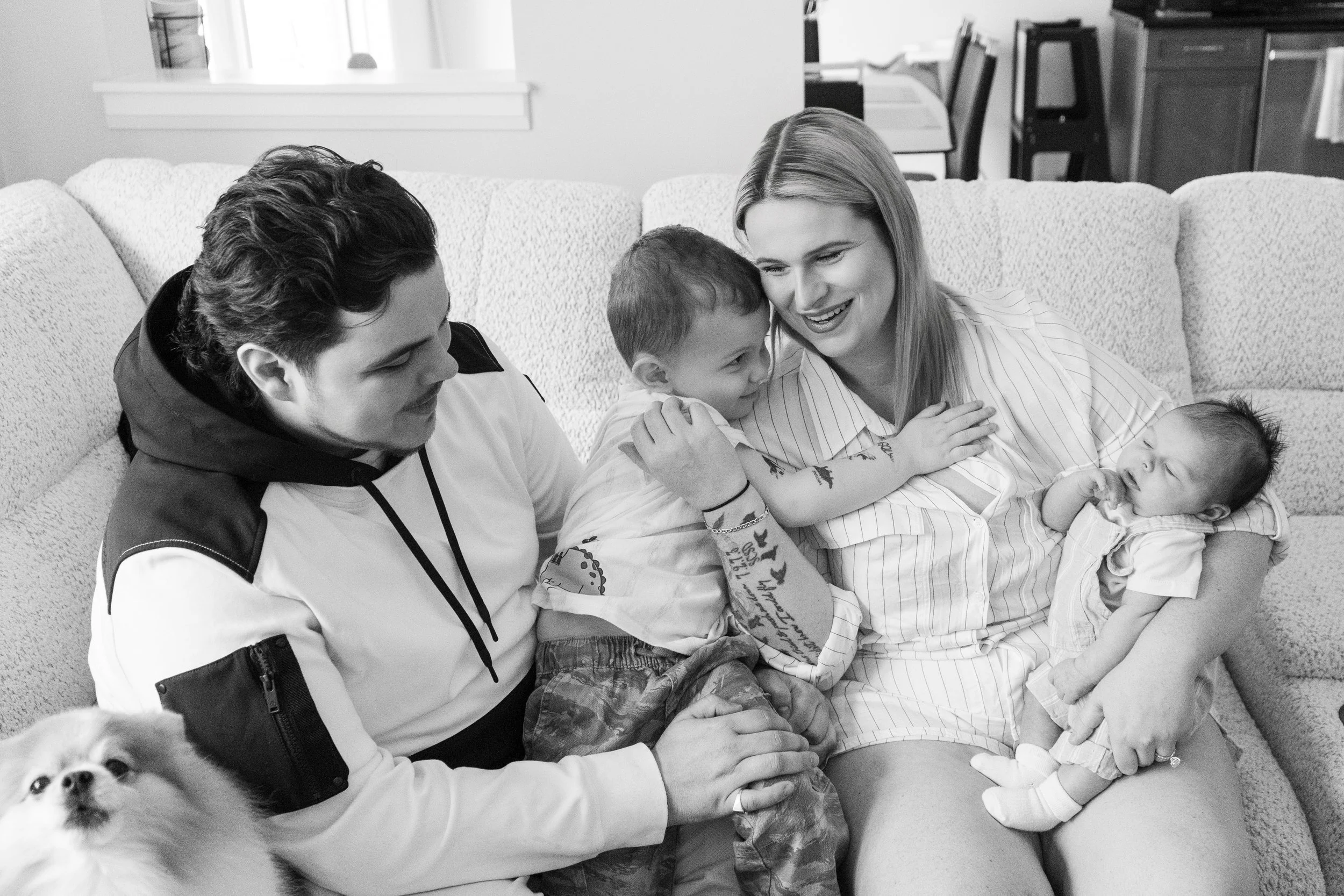 A family sitting on a couch, with a woman and a young boy hugging a sleeping baby, and a dog looking up at the camera.