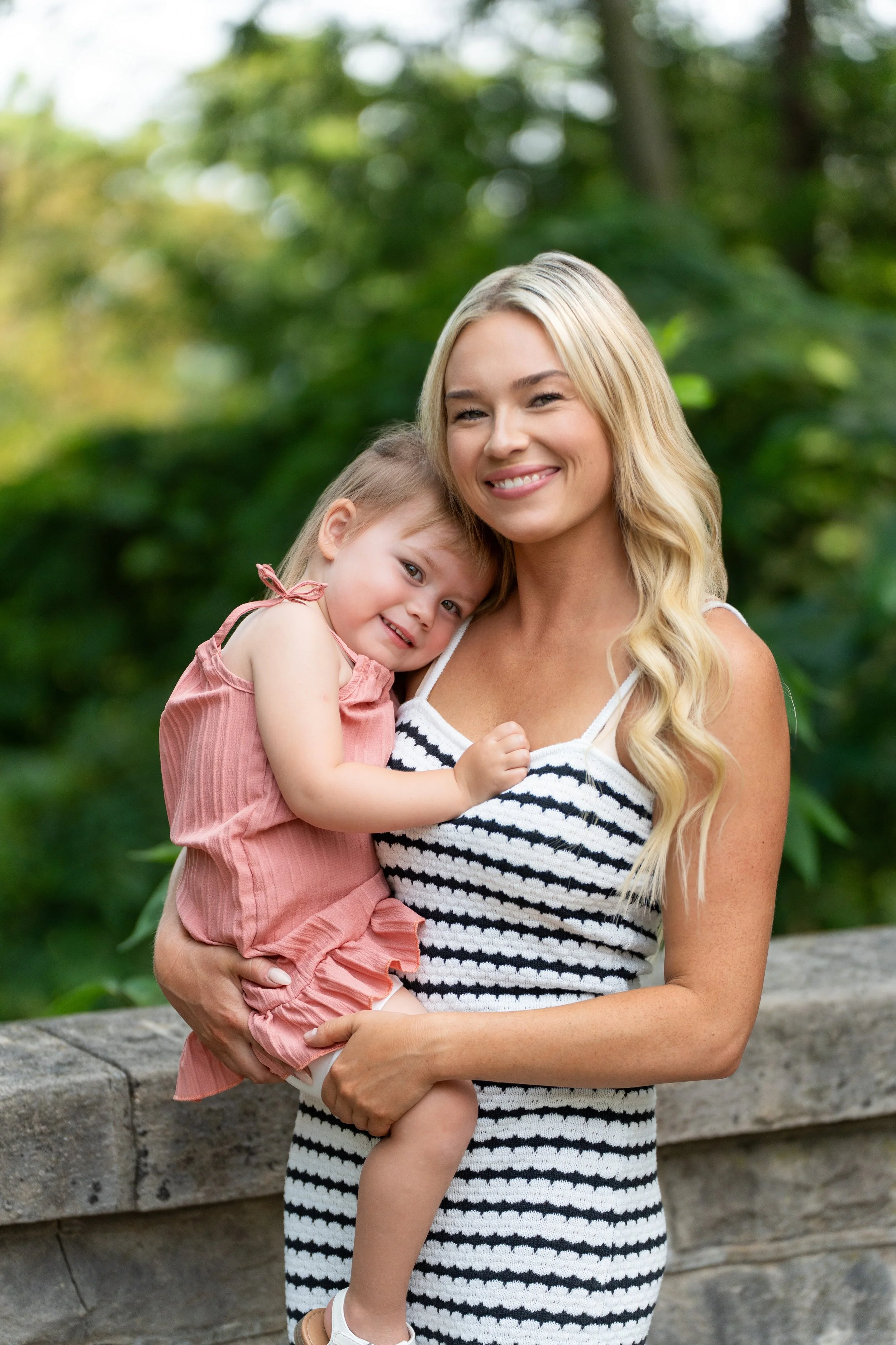 A woman with long blonde hair, wearing a black and white striped dress, holding a smiling young girl in a pink dress with ruffles, outdoors in a lush green environment.