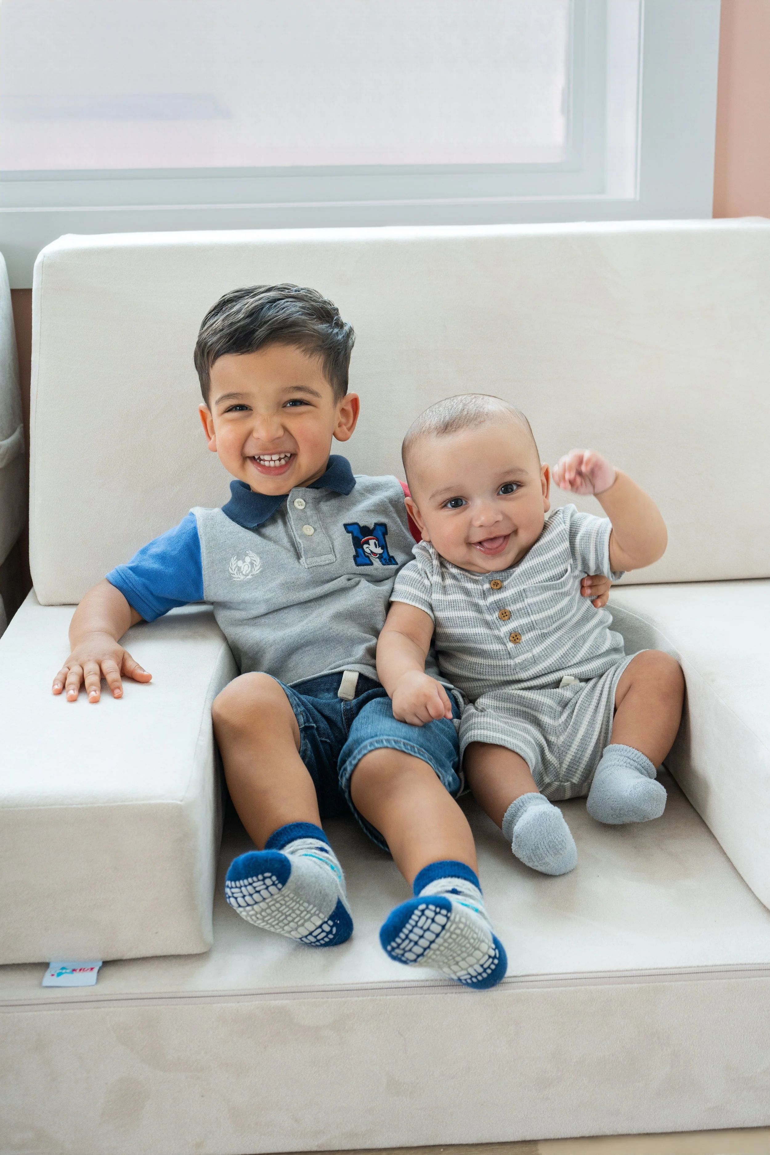 Two young boys sitting on a white couch, smiling and appearing happy. The older boy has dark hair and is wearing a gray and blue polo shirt, while the younger boy has light hair and is wearing a gray and white striped outfit. They both have socks wit