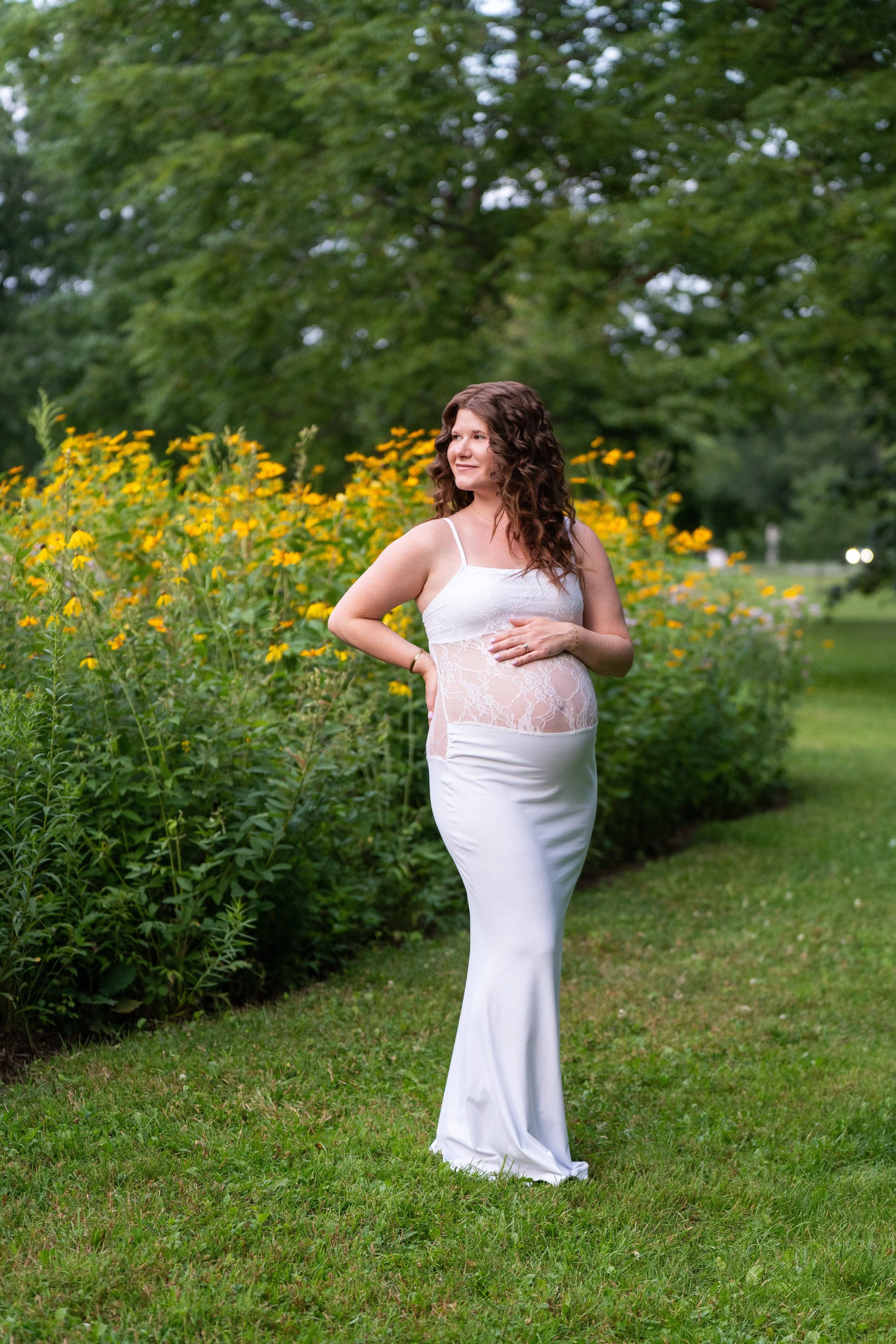 A woman in a white dress standing outdoors in a garden with yellow flowers and green trees in the background.