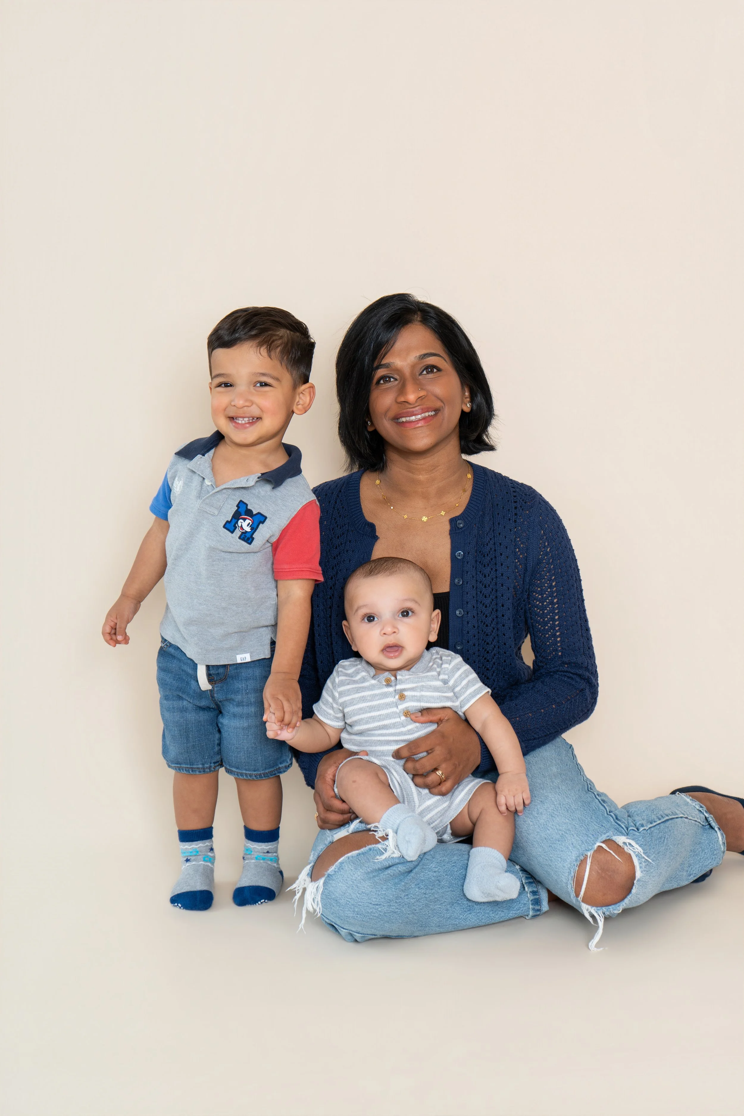 A woman with short black hair and wearing a navy blue cardigan is sitting on the floor, holding a baby with light-colored hair and a striped shirt. A young boy with short dark hair, gray and red shirt, and denim shorts is standing next to her, holdin