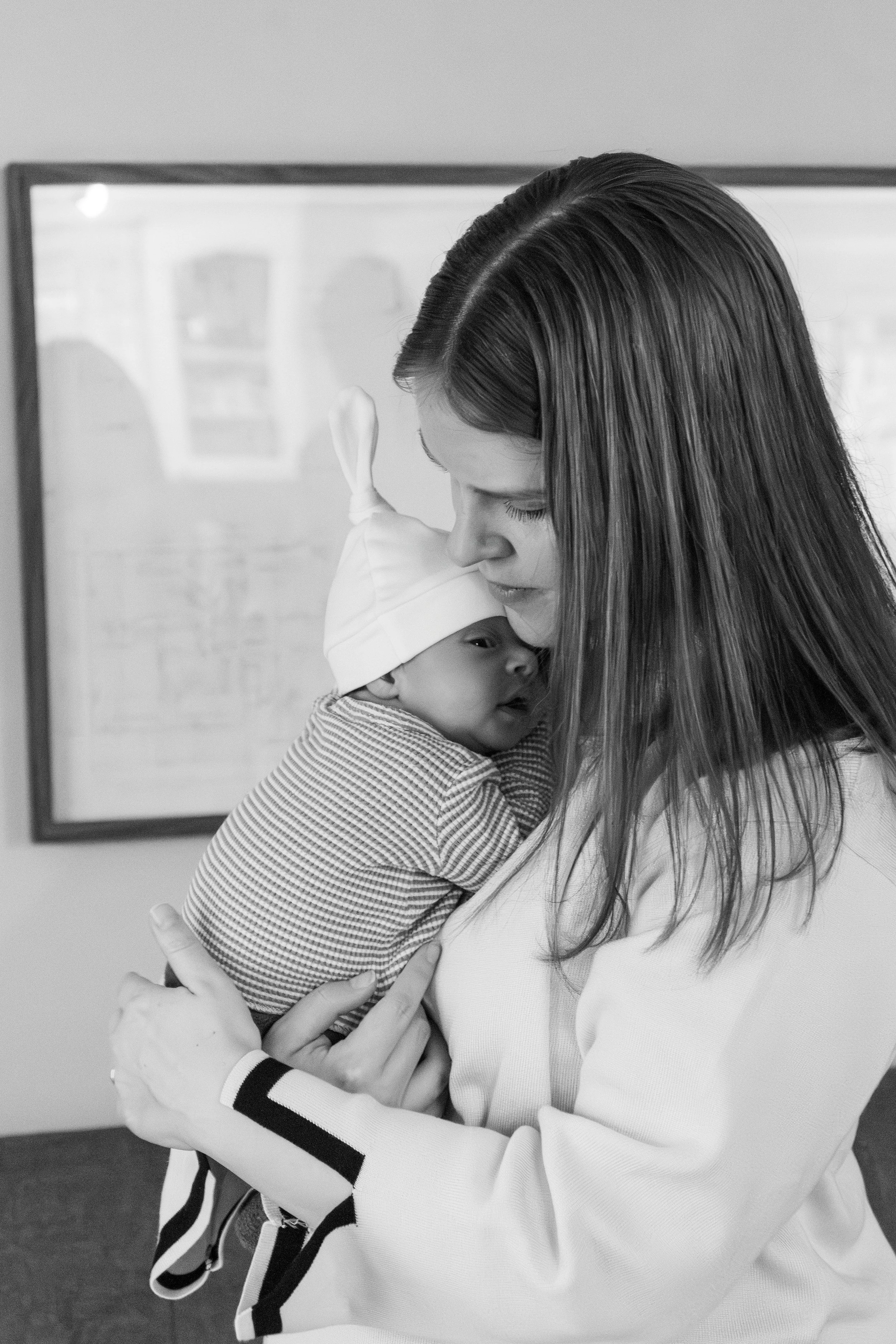 A woman with long, dark hair holding a young child who is wearing a white cap and striped shirt, inside a room with a framed picture on the wall.