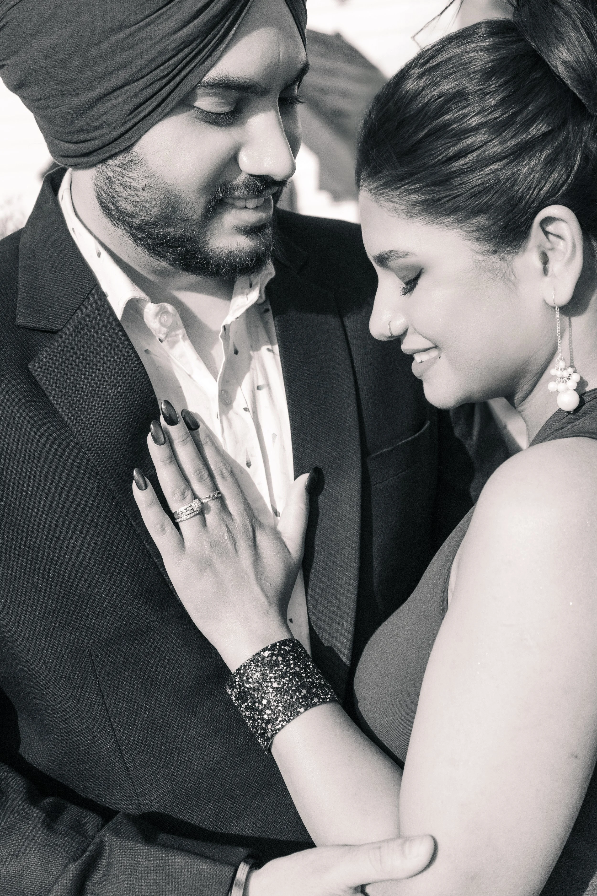 A black and white photograph of a man and woman close together, smiling with foreheads touching. The man is wearing a turban and a suit, while the woman has earrings and a tattoo on her arm. They appear to be enjoying a joyful moment.