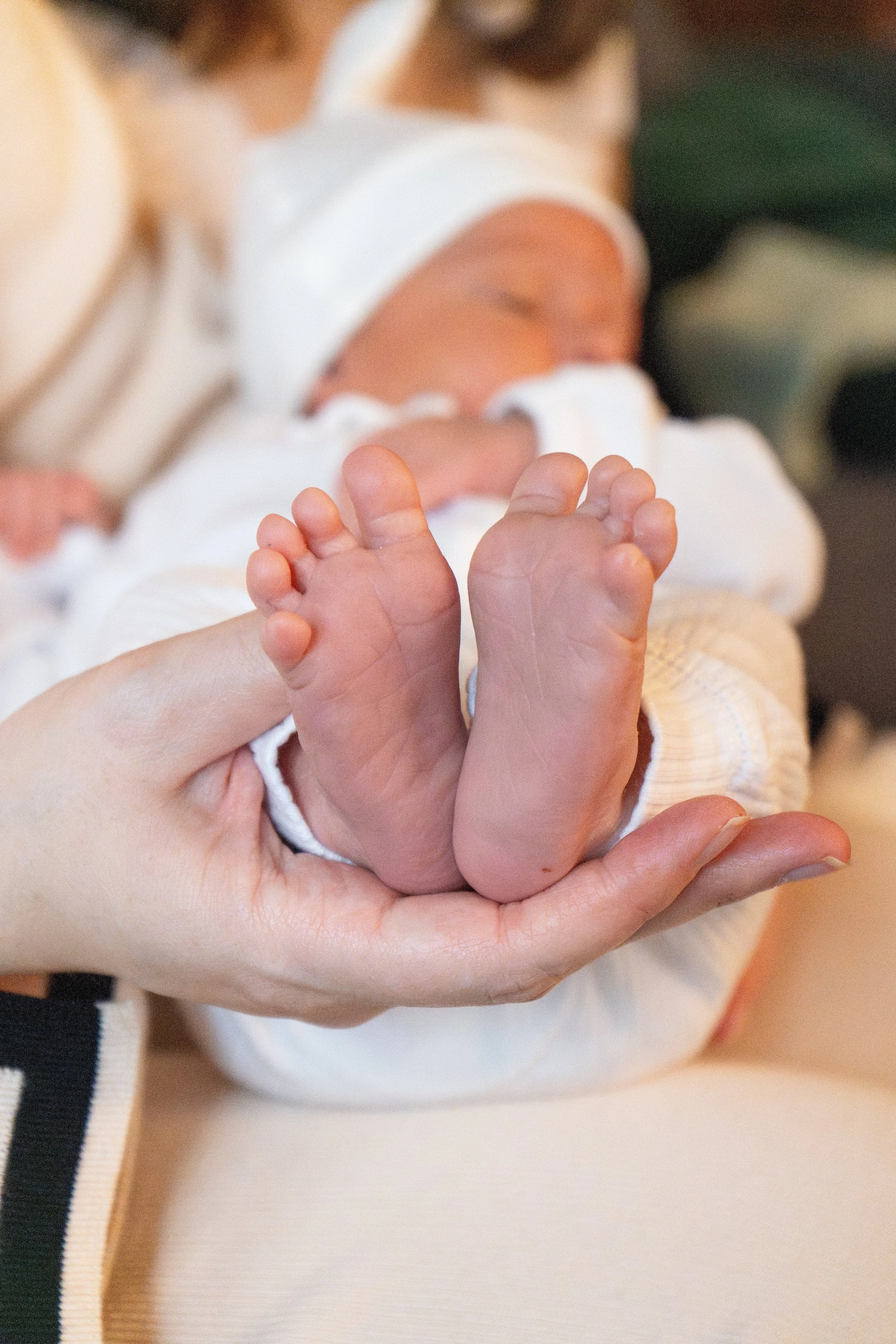 Close-up of an adult holding a baby's feet with both hands, baby dressed in white, background blurred.