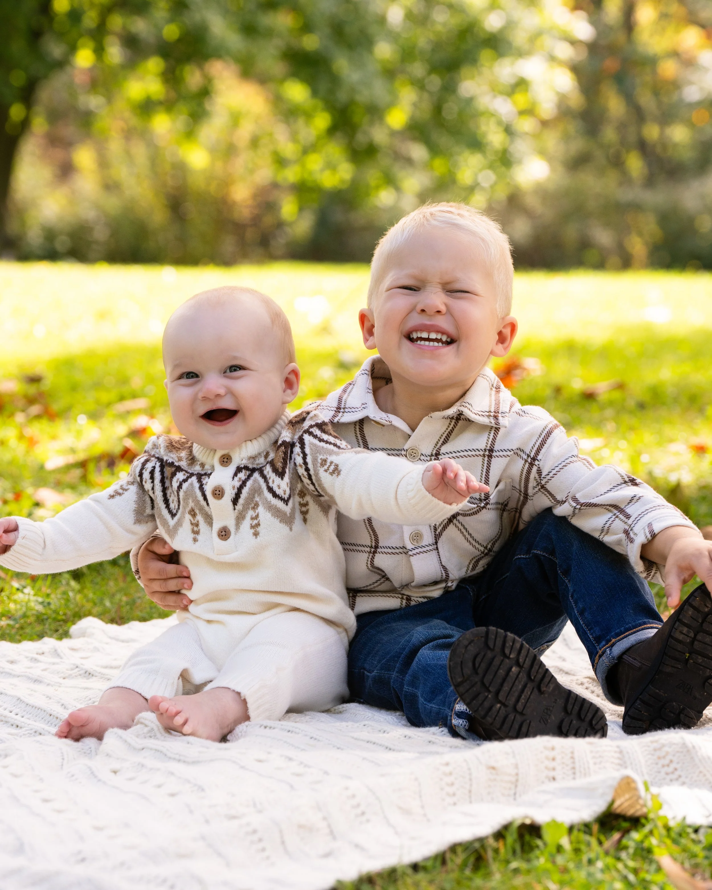 Two young boys sitting on a blanket outdoors in a park, smiling and enjoying the sunshine with green trees and grass in the background.