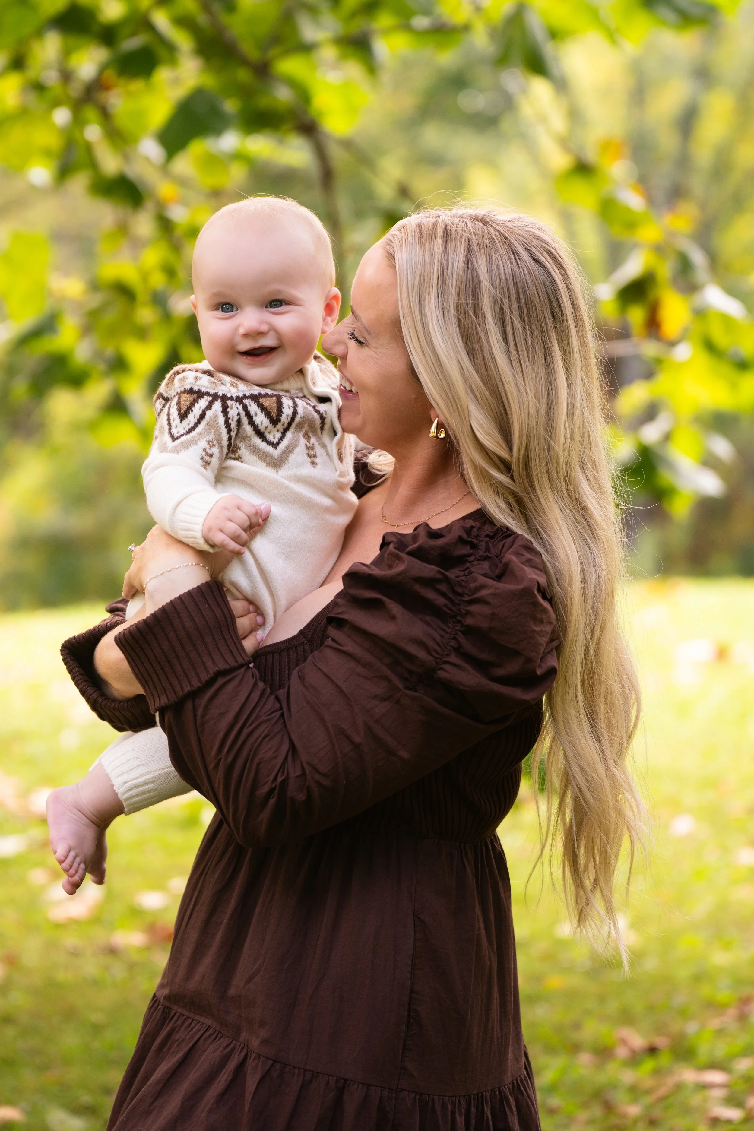 A woman holding a smiling baby outdoors in a park with green trees and grass.