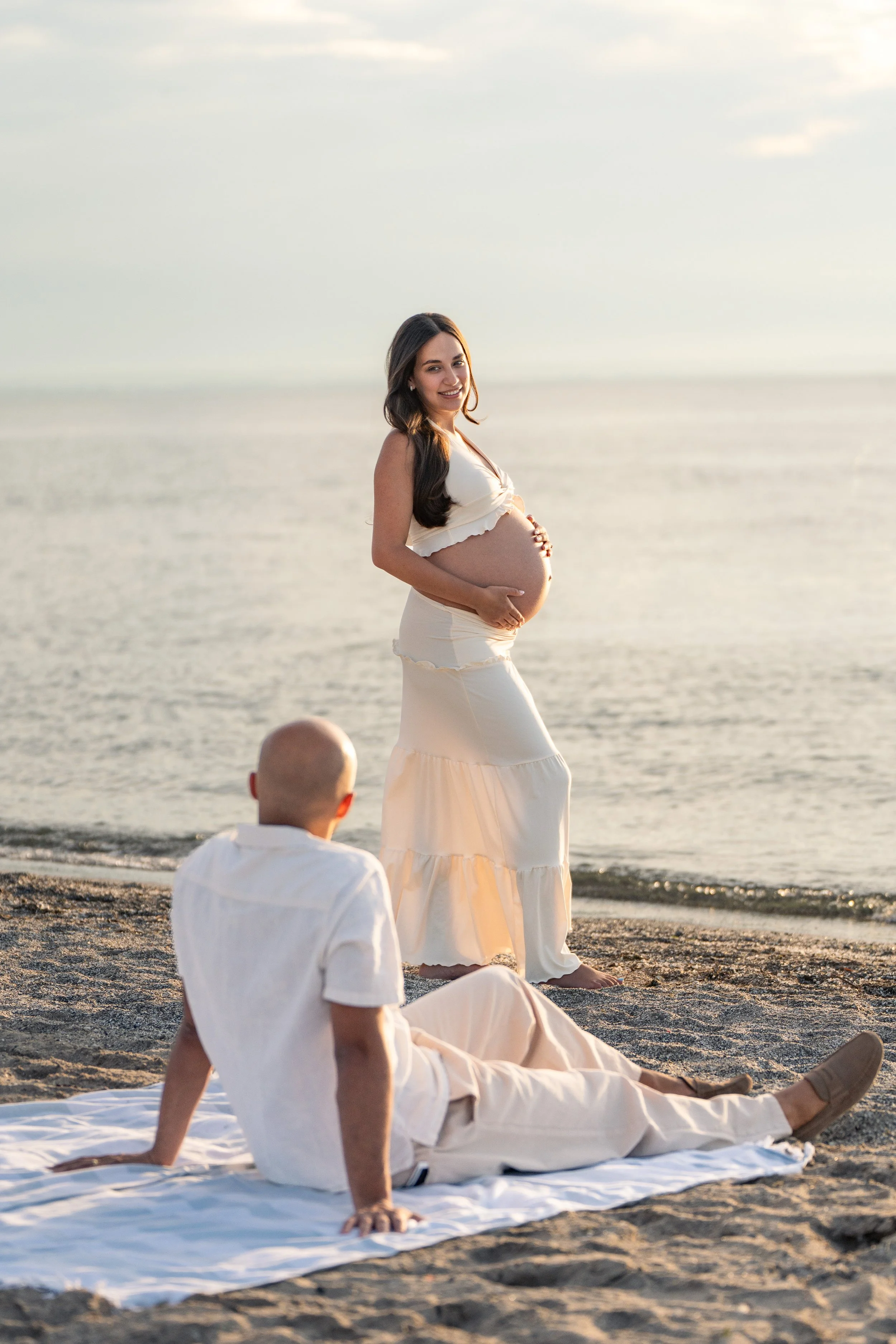 A pregnant woman with long brown hair cradling her belly on Sunset beach in St. Catharines while looking over at her partner at sunset.