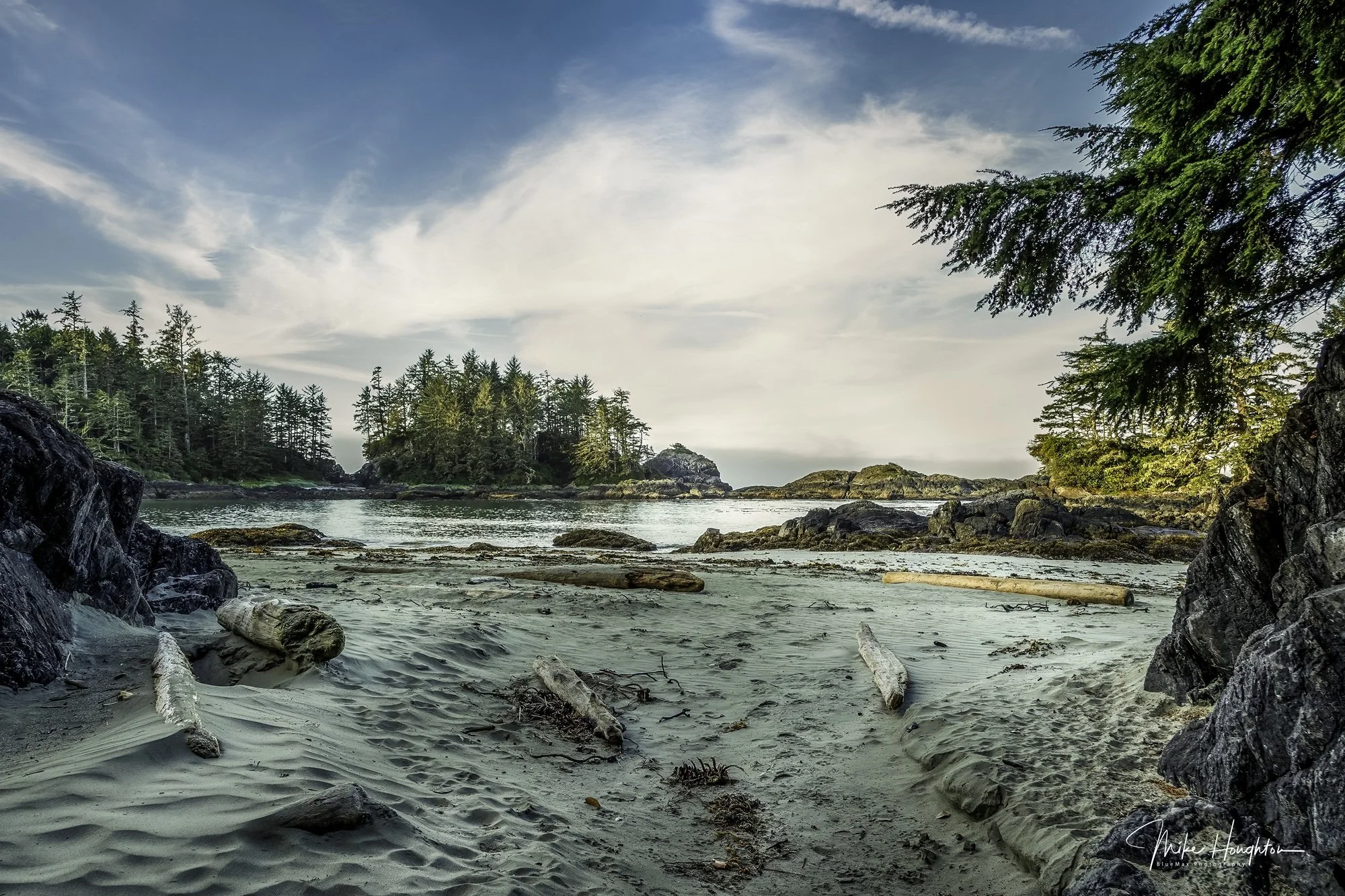 A peaceful coastal scene with sandy beach, driftwood, rocky formations, and green trees lining the shore under a blue sky with wispy clouds.