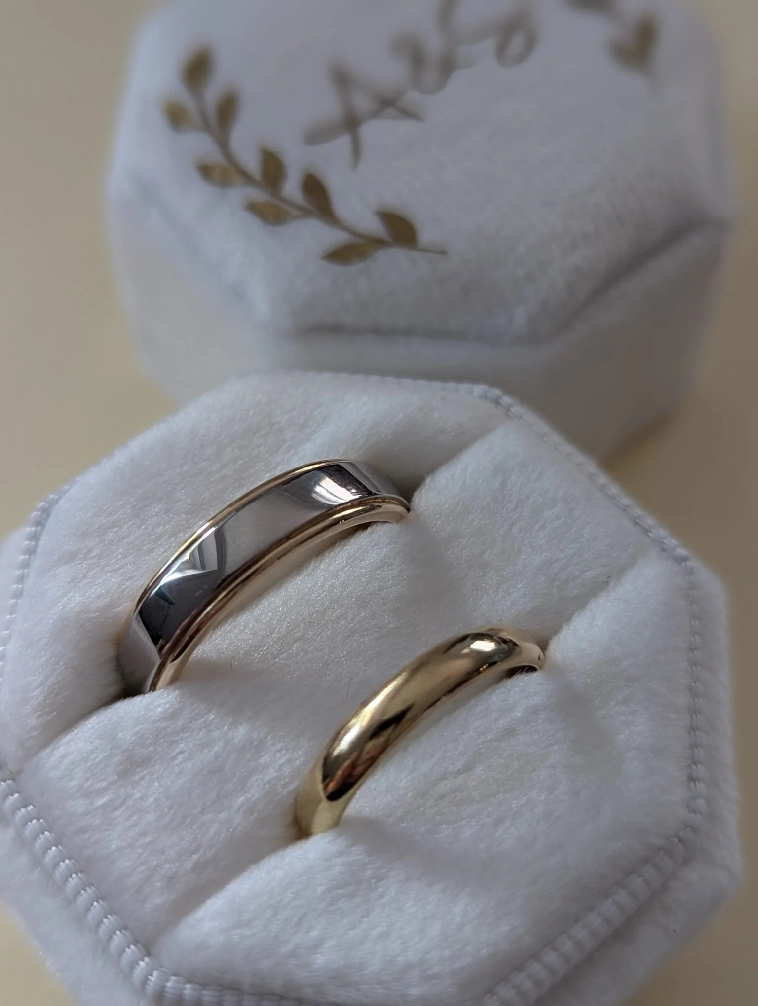 Two wedding bands in a white velvet ring box, one silver and one gold, with a white cloth background.