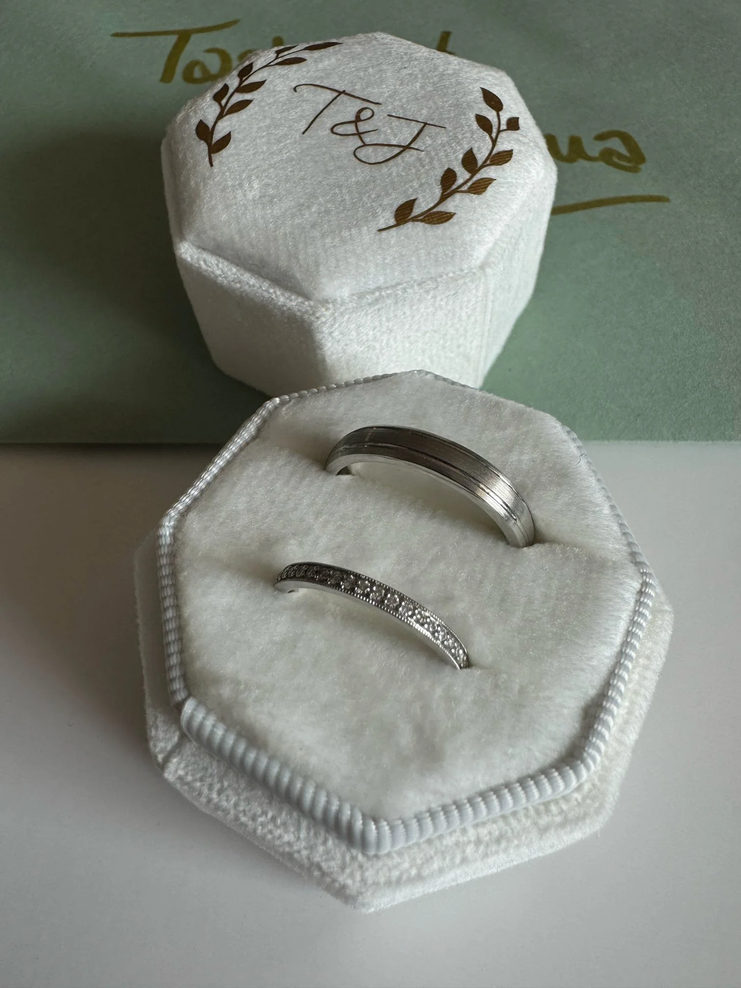 A jewelry box with two rings inside, one plain band and one with small stones, and a white cover with gold lettering and decoration in the background.
