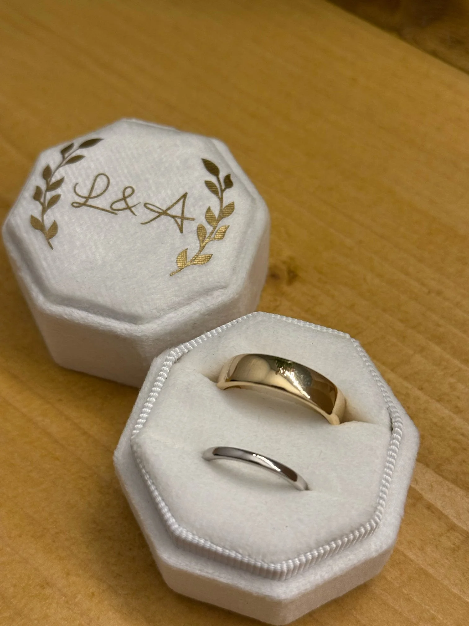 A pair of wedding rings, one gold and one silver, inside a white, hexagonal ring box with a cushioned interior. The box with gold rings has a lid with gold lettering and laurel designs that appears to spell 'L&A', placed on a wooden surface.