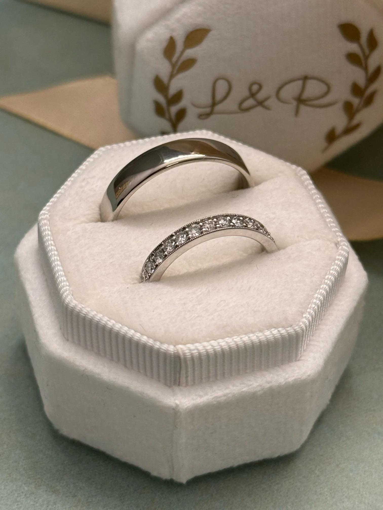 Two wedding rings, one plain and one with embedded diamonds, in a white ring box with a soft interior, with a decorative box in the background.