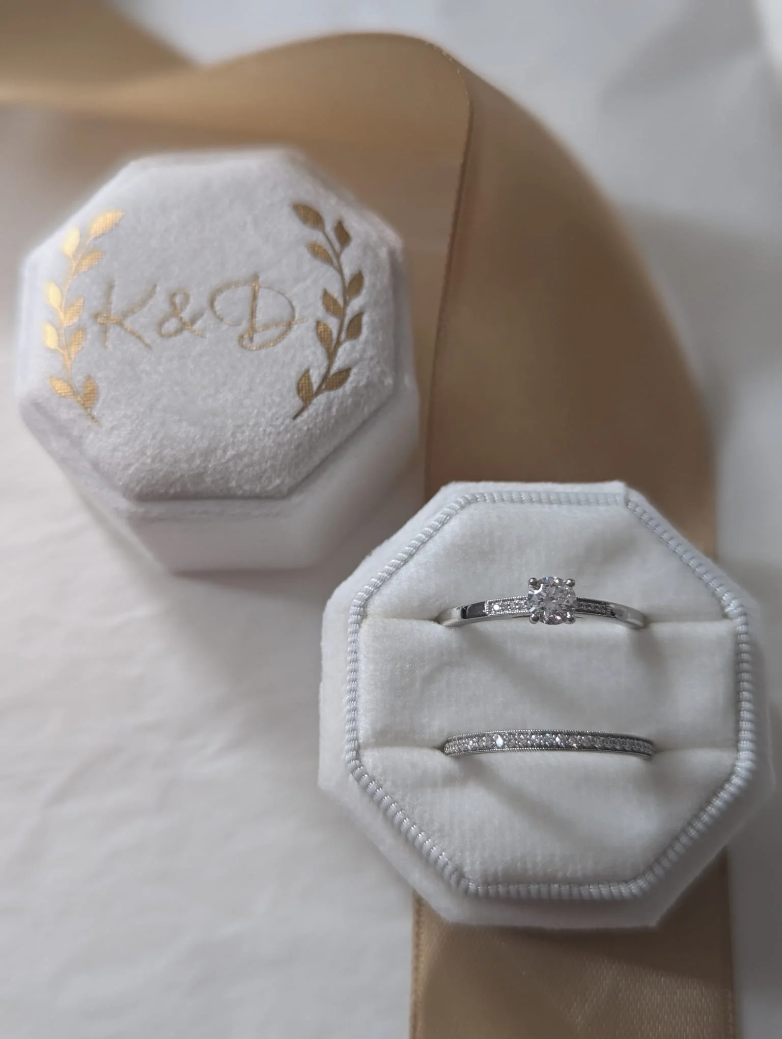 Close-up of a white jewelry box with diamond rings inside, beside a container with initials "K&D" and gold laurel leaf design, set on beige fabric.