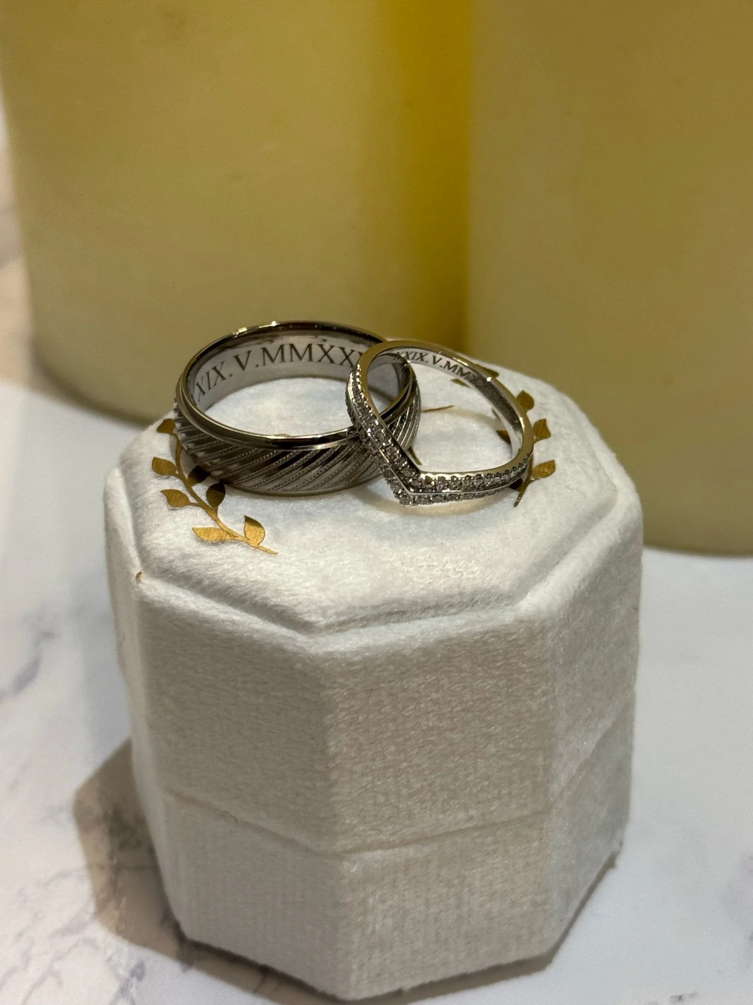 Two wedding rings, one with engraved Roman numerals inside and the other with small diamonds, placed on a white textured ring box with gold leaf decorations.