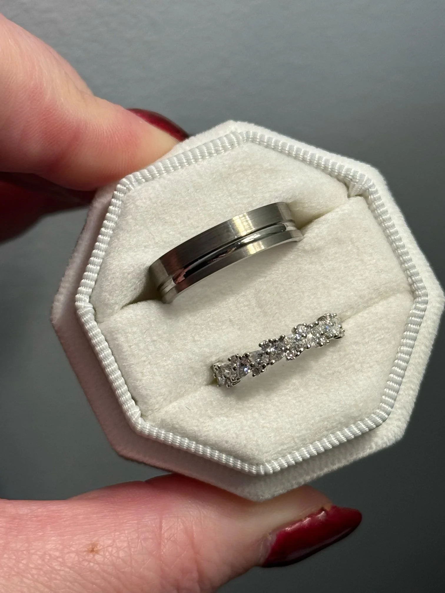 A jewelry ring set inside a white, padded ring box held by a person with red-painted nails. The set includes a plain metallic band and a diamond-studded band.
