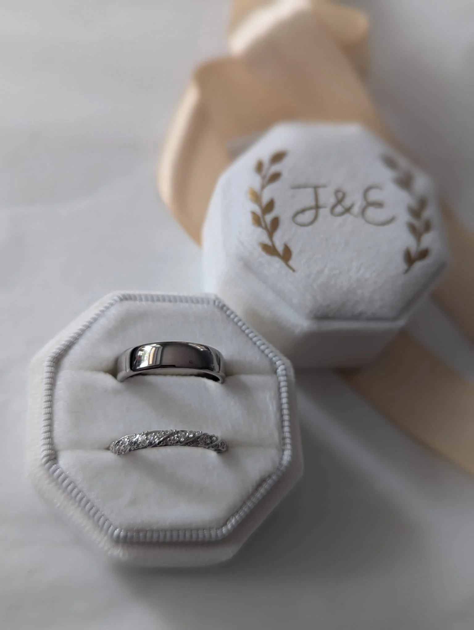 Two rings, one plain band and one with diamonds, inside a white ring box, with a wedding cake in the background with the initials J & E and gold leaf accents.