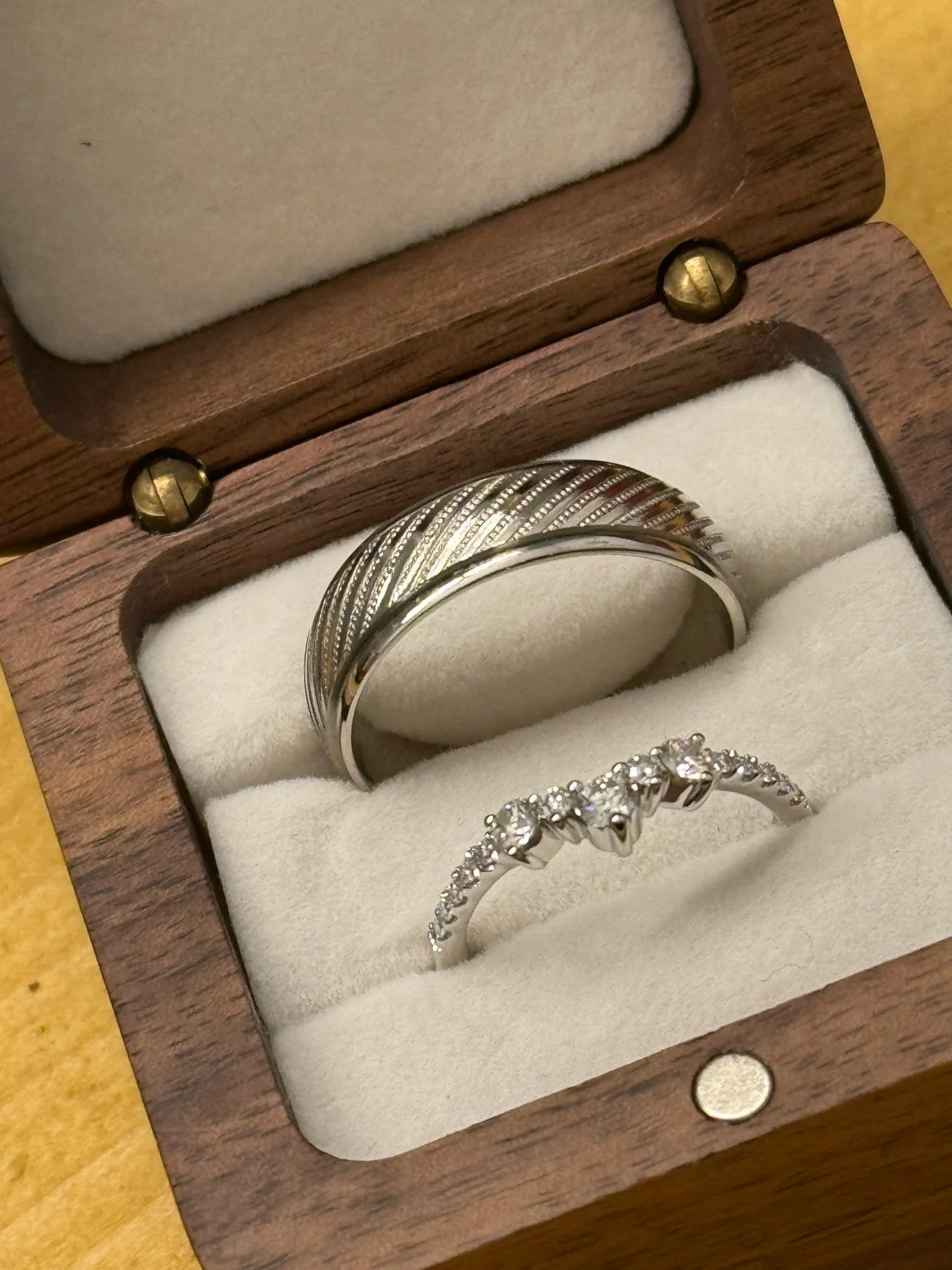 A wooden ring box containing a silver wedding band and an engagement ring with small diamonds.