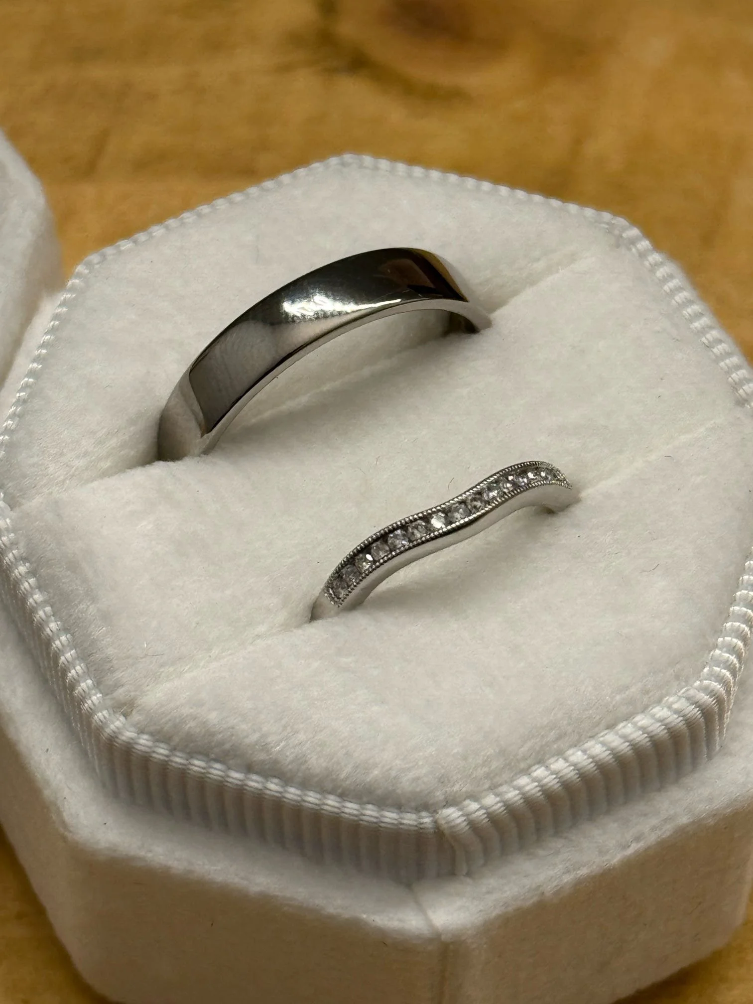 A pair of rings in a white ring box, one plain silver band and one with diamonds.