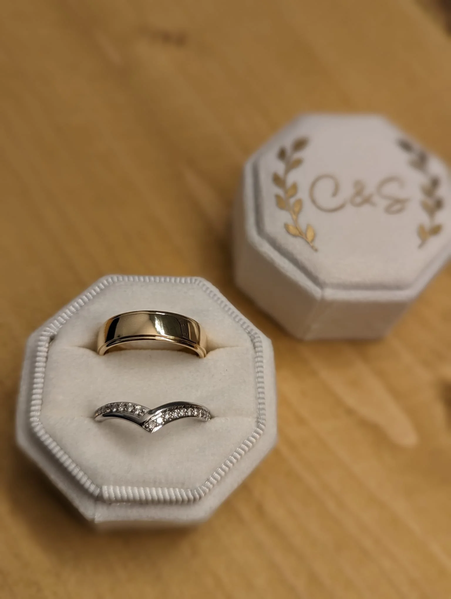 A white jewelry box with gold detailing contains two rings: a gold band and a silver ring with small diamonds. The box is on a wooden surface.