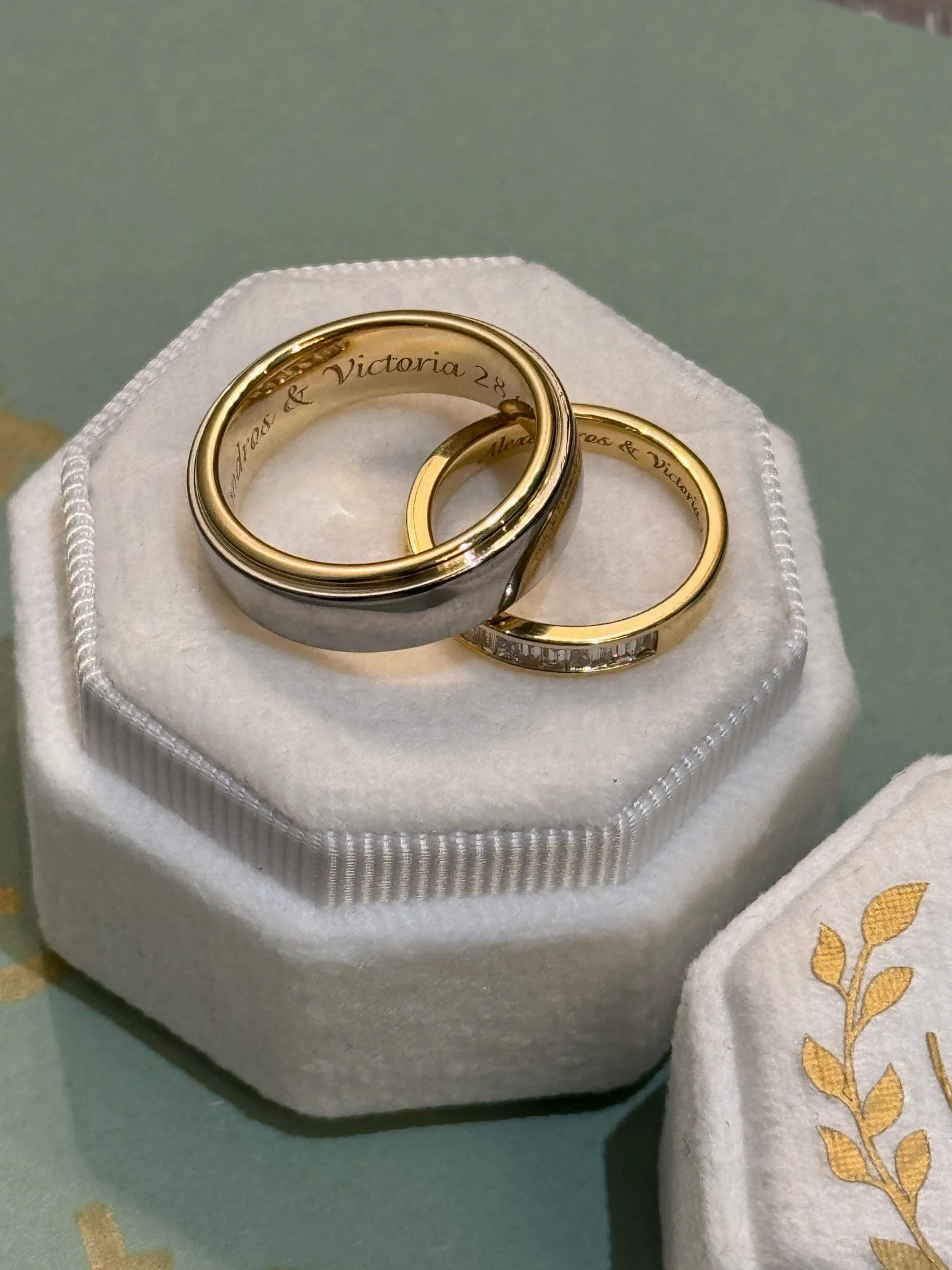 Two wedding rings, one gold with engraved text inside, and the other a gold band with small diamonds, displayed on a white, velvet ring box.