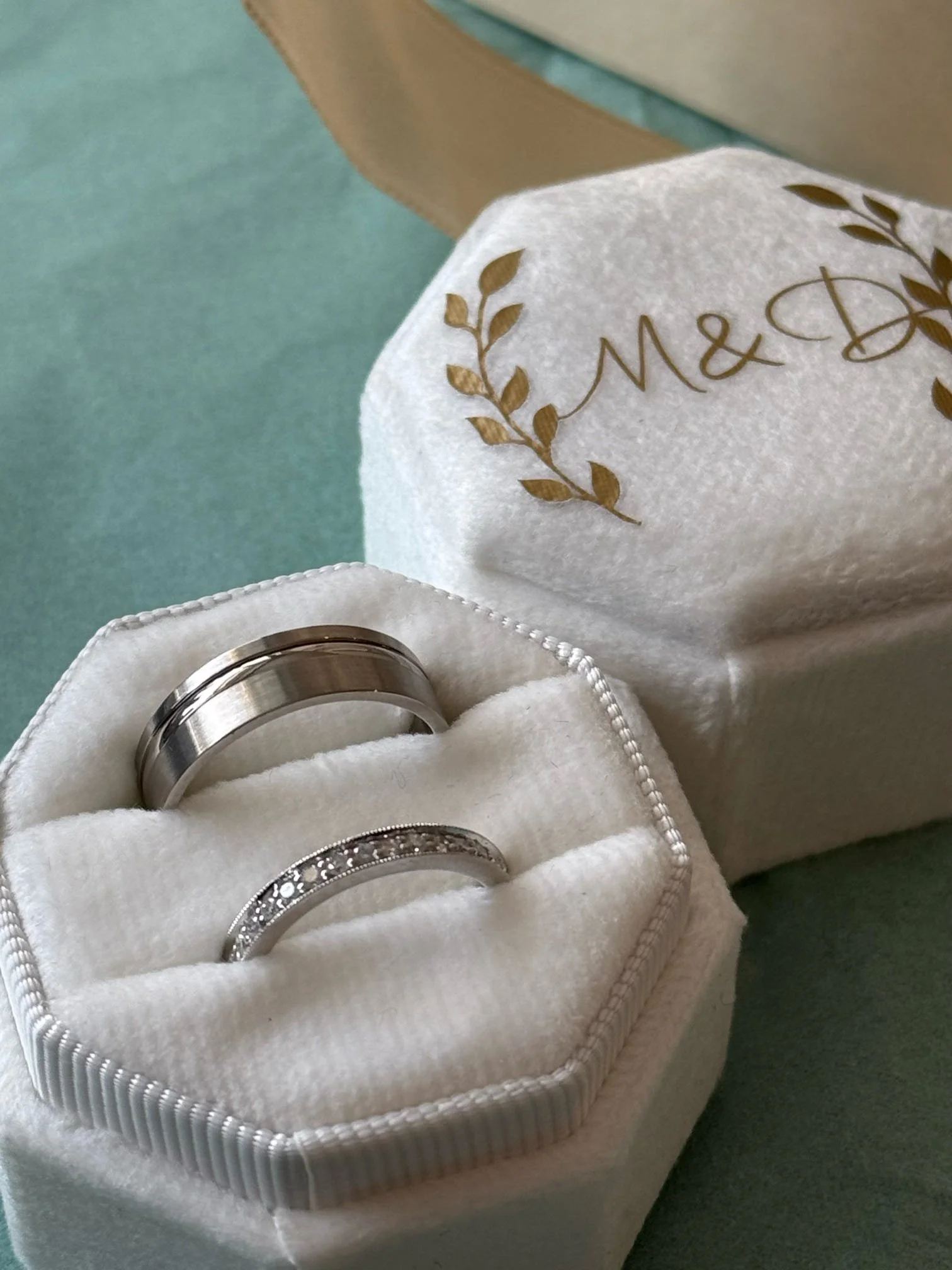 A jewelry box with two wedding rings inside, one plain silver band and one silver band with small stones, on a green surface. The jewelry box lid, marked with gold writing and decorative leaves, is partially open.