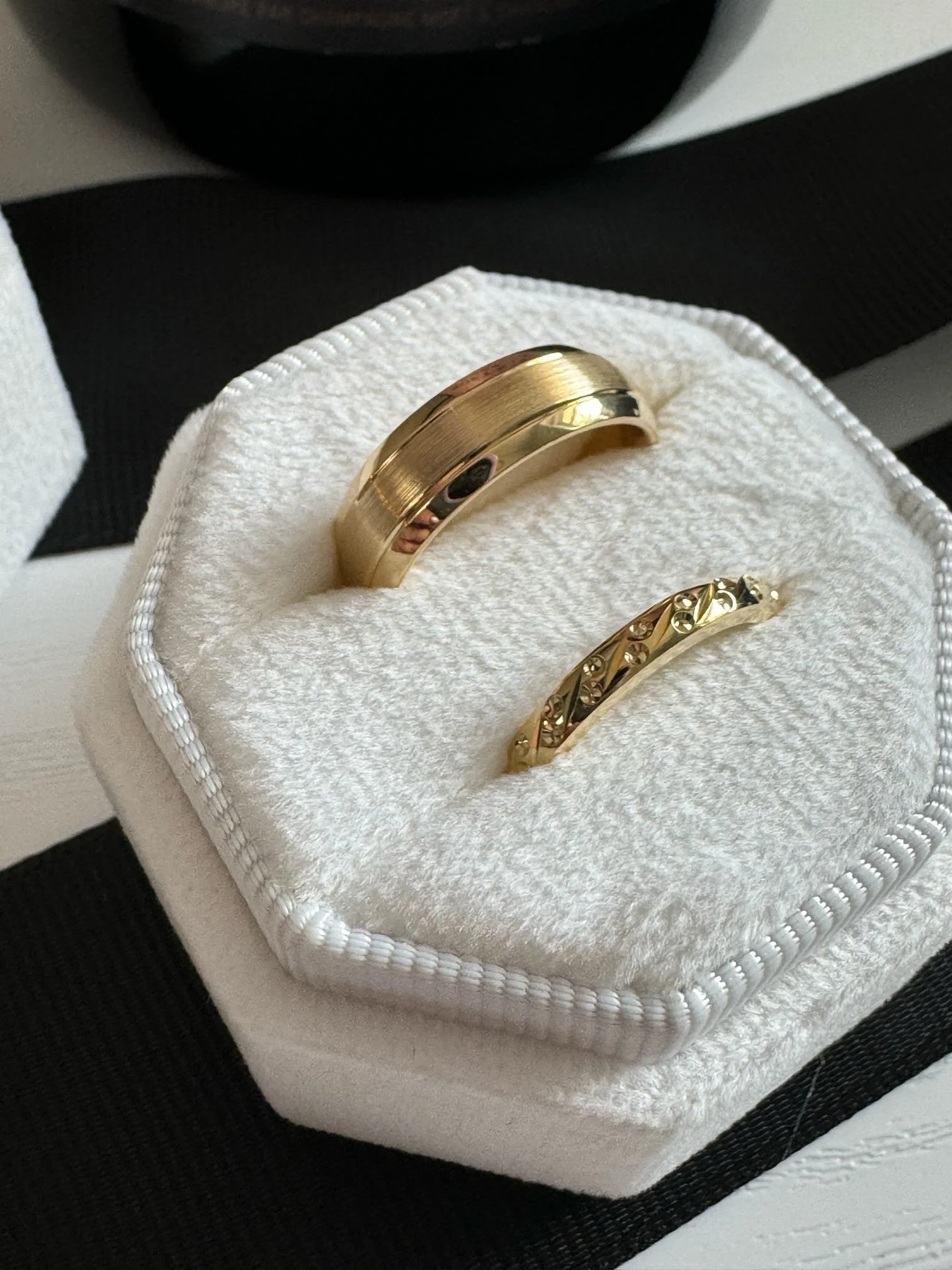 Gold wedding bands, one plain and one with decorative design, in a white jewelry box with black ribbon.