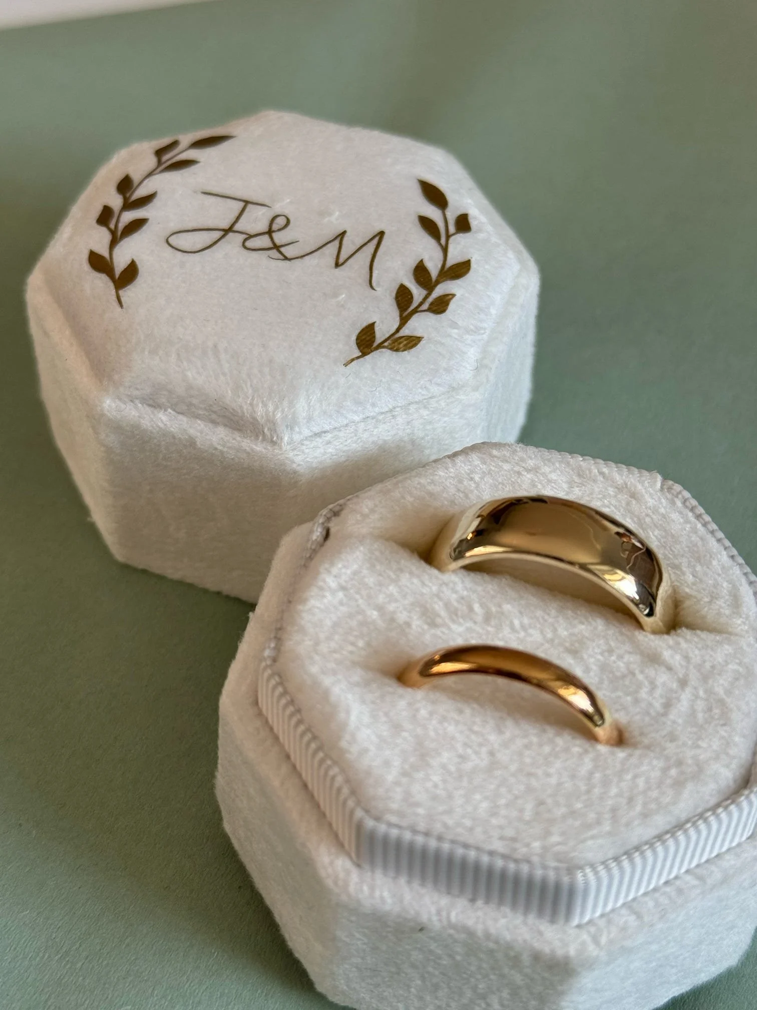 Gold wedding bands in a white velvet box with a matching cover with 'J&M' and a laurel design.