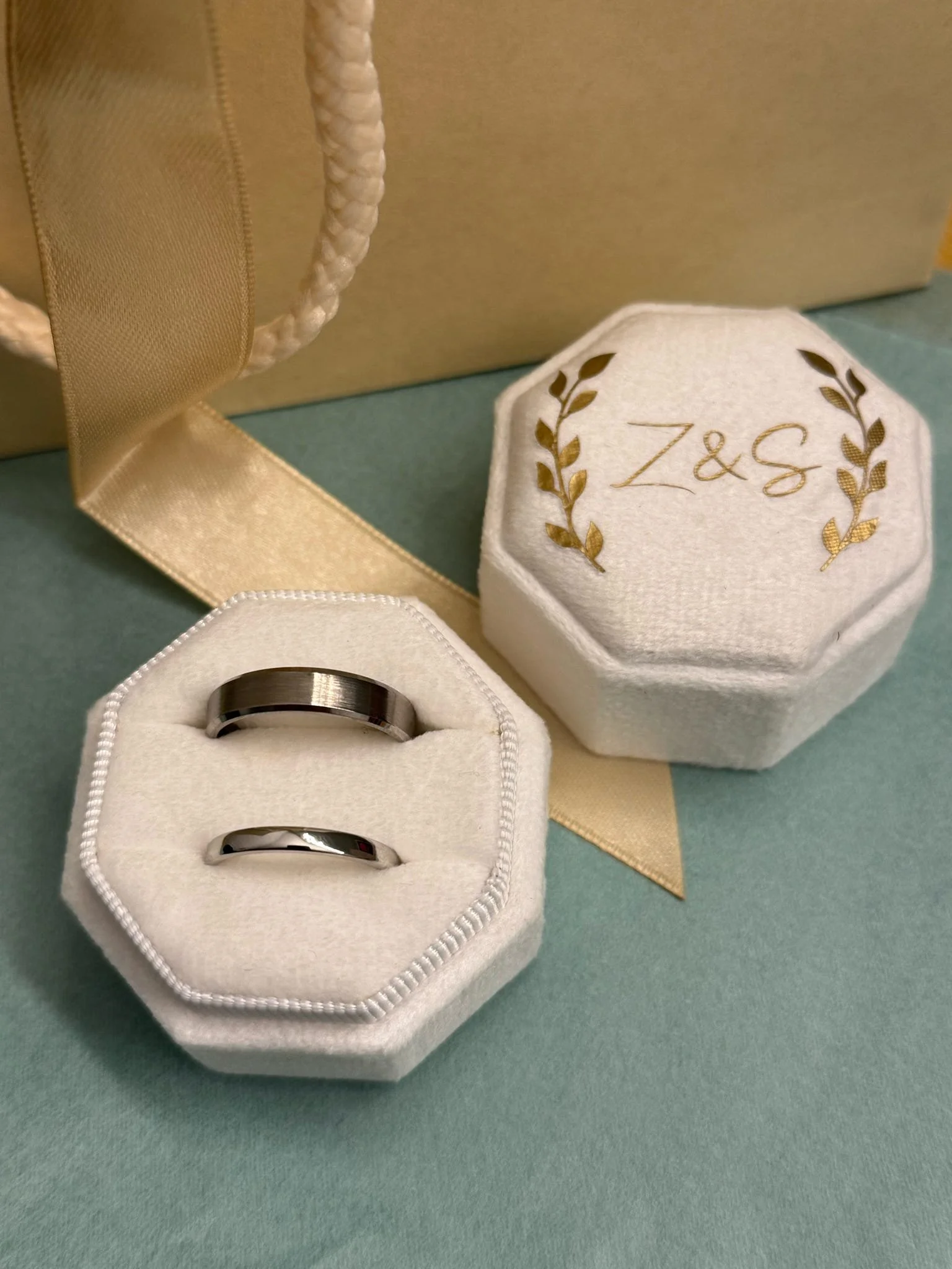 Two wedding bands resting inside white velvet ring box, with another box beside it embroidered with 'Z & S' surrounded by gold leaves, on a gold and turquoise surface with a silk ribbon.
