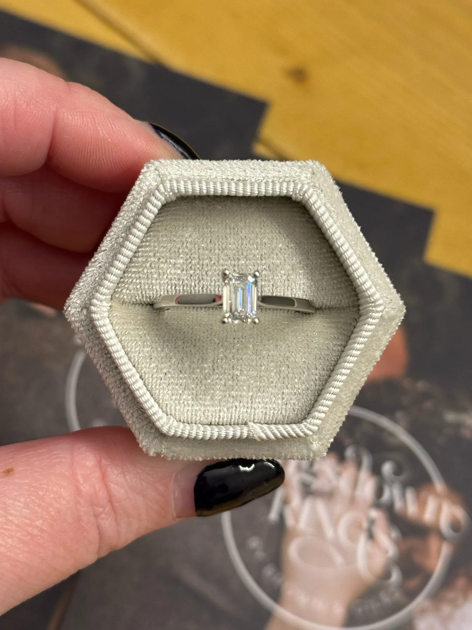 A silver ring with an emerald-cut clear gemstone, possibly a diamond, in a beige velvet ring box.