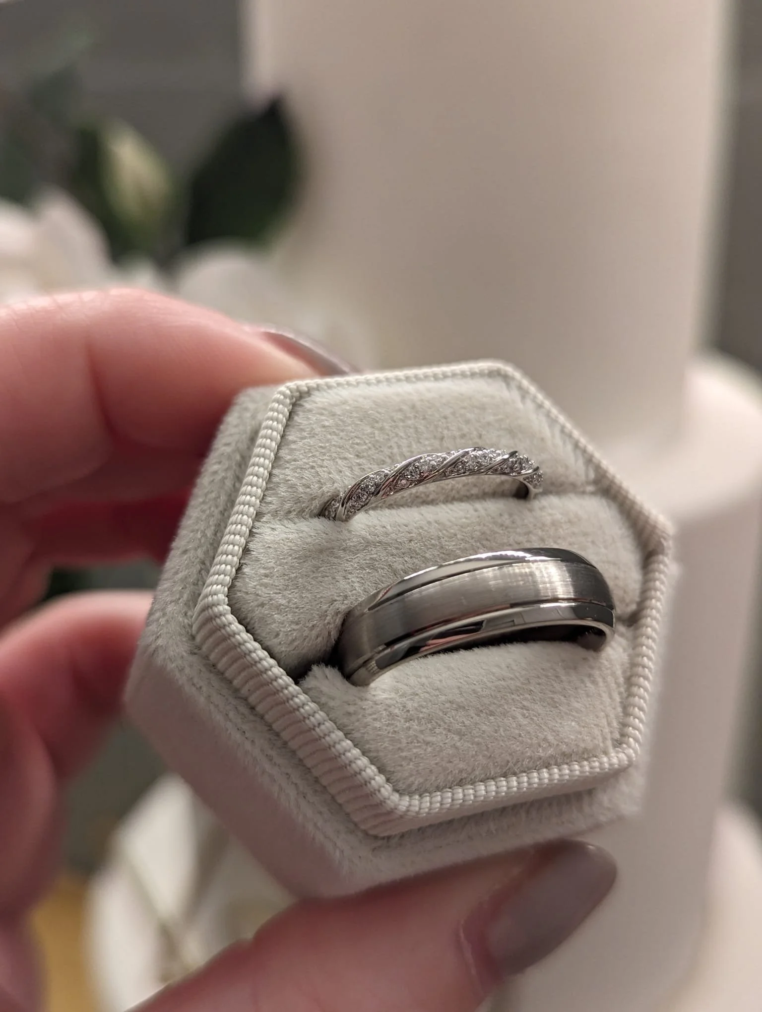 A hand holding a beige ring box with two silver rings inside, one with a twisted design and small stones, and the other with a smooth, wide band.