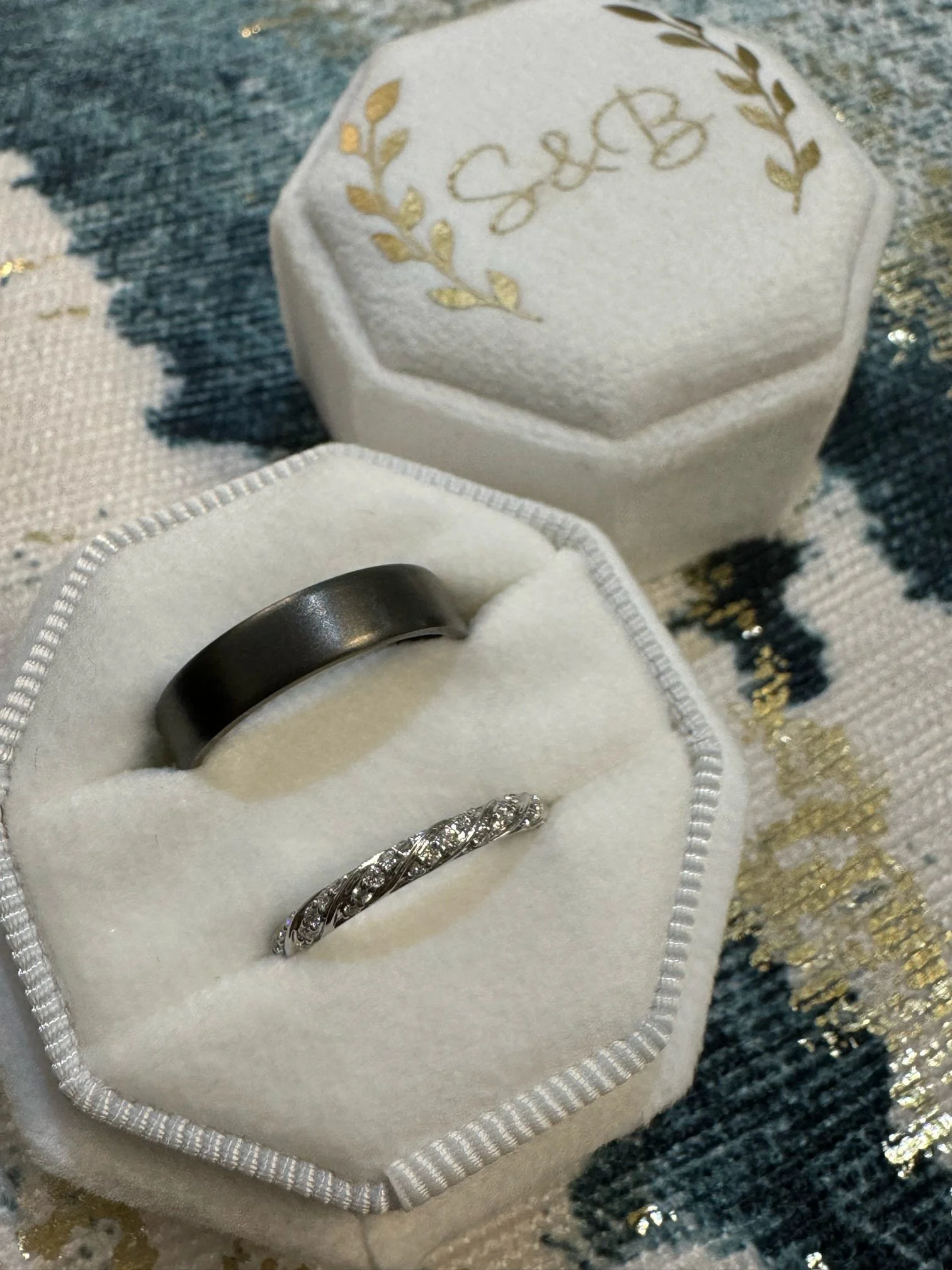 A pair of wedding rings in a white velvet ring box, one black band and one diamond-studded band, on a patterned fabric surface. The lid of the box has gold lettering and a decorative laurel wreath design.