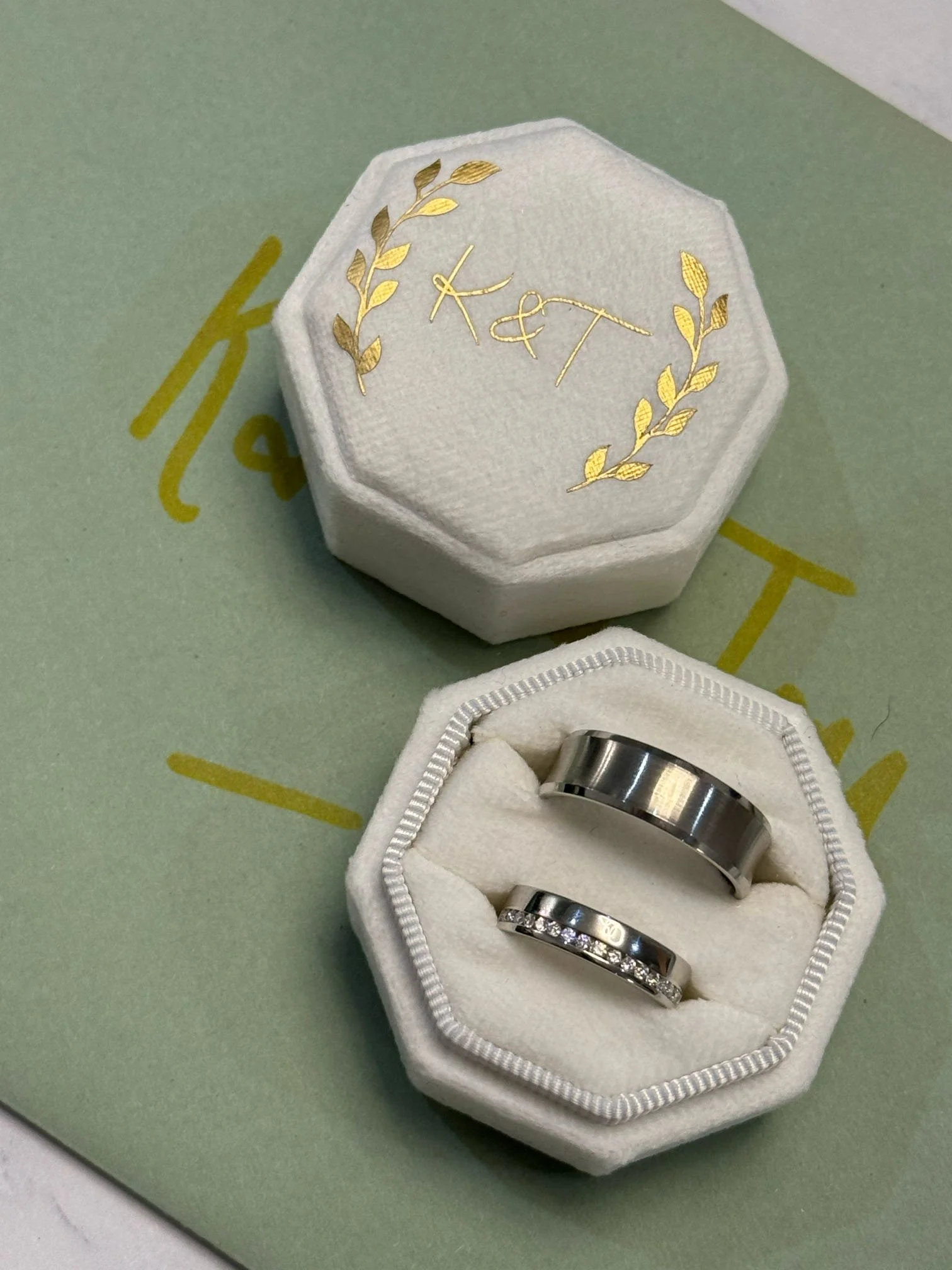 A pair of wedding rings in a white velvet ring box with gold lettering and decorative leaves on the lid, placed on a green surface with partially visible gold text.