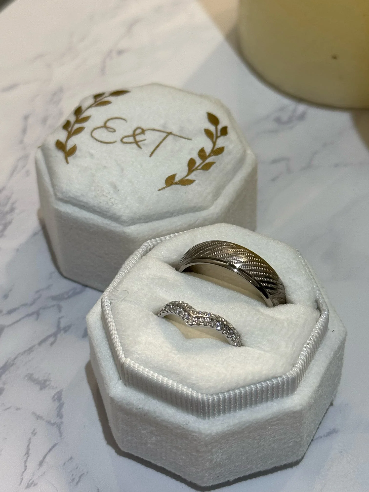 Wedding rings in a white padded box with gold initials and laurel design on the lid, placed on a marble surface.