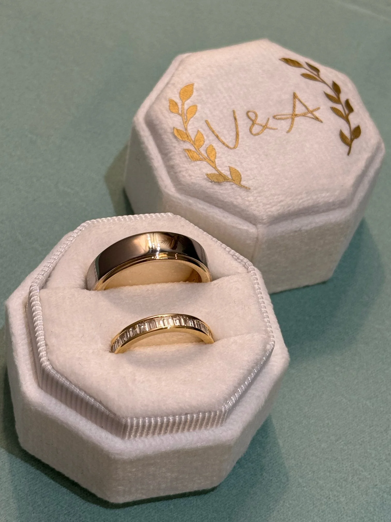 Two rings in a white ring box with a matching lid, both rings are gold, one with a smooth band and the other with embedded rectangular gems. The box is open and sitting on a green surface, with the lid placed behind it. The lid has the word 'Love' embroidered in gold along with decorative gold leaves.