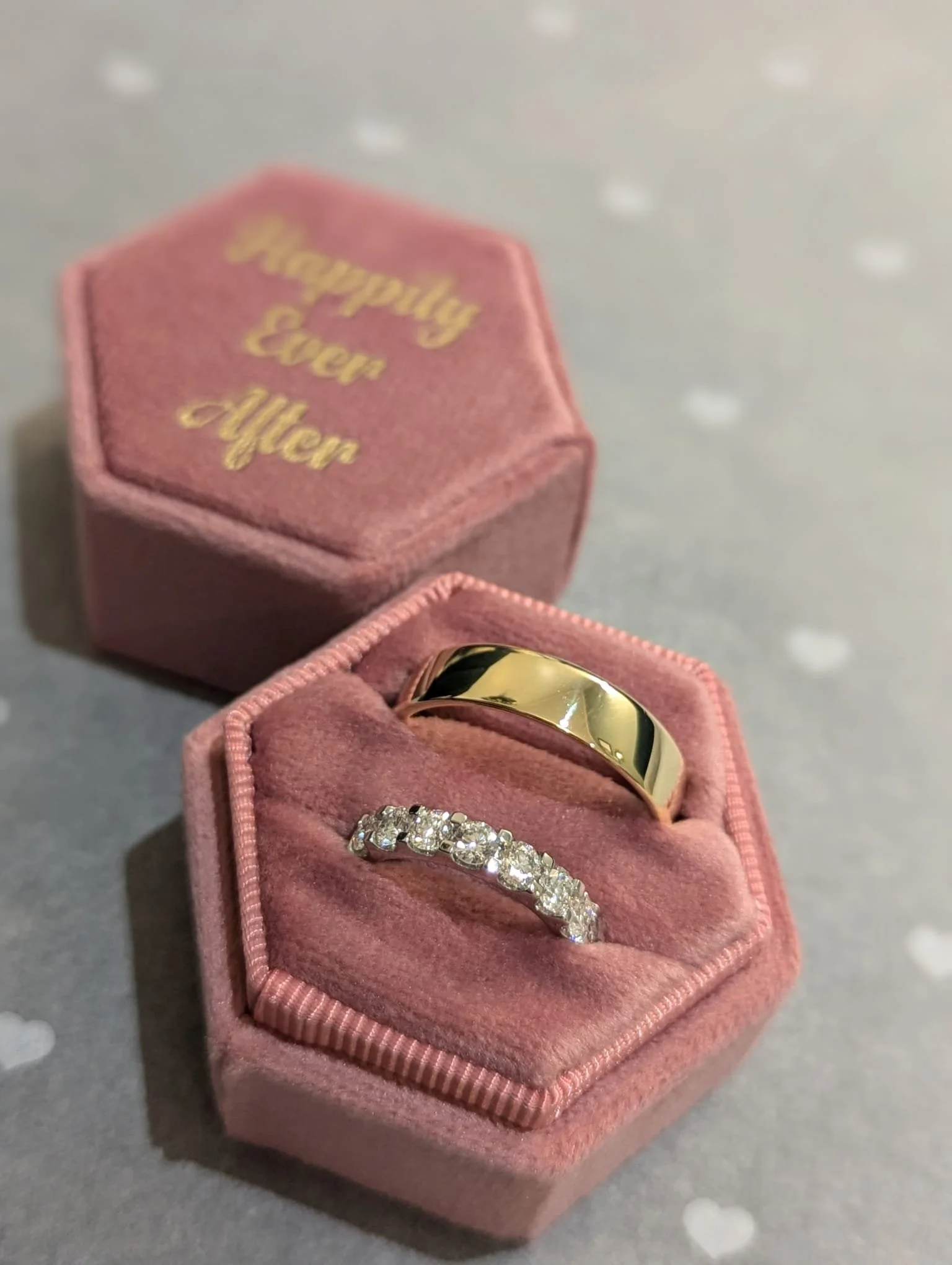 Pink jewelry box with gold rings inside, one is plain gold and the other is band with diamonds. The box lid is open and has gold lettering that reads 'Happily Ever After.'