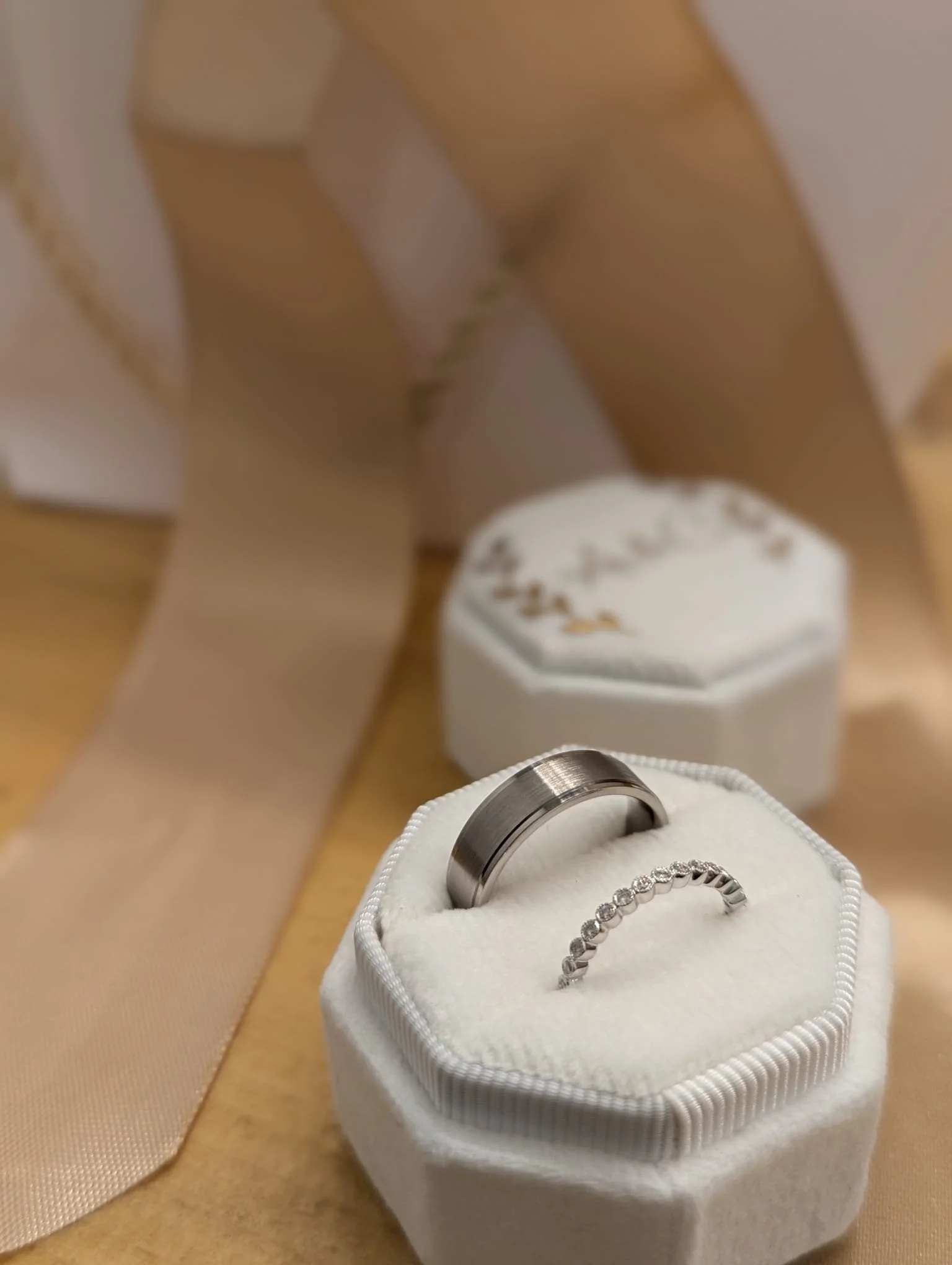 Two silver rings, one plain band and one with embedded stones, displayed on white ring box with ribbon, surrounded by decorative boxes and ribbon.