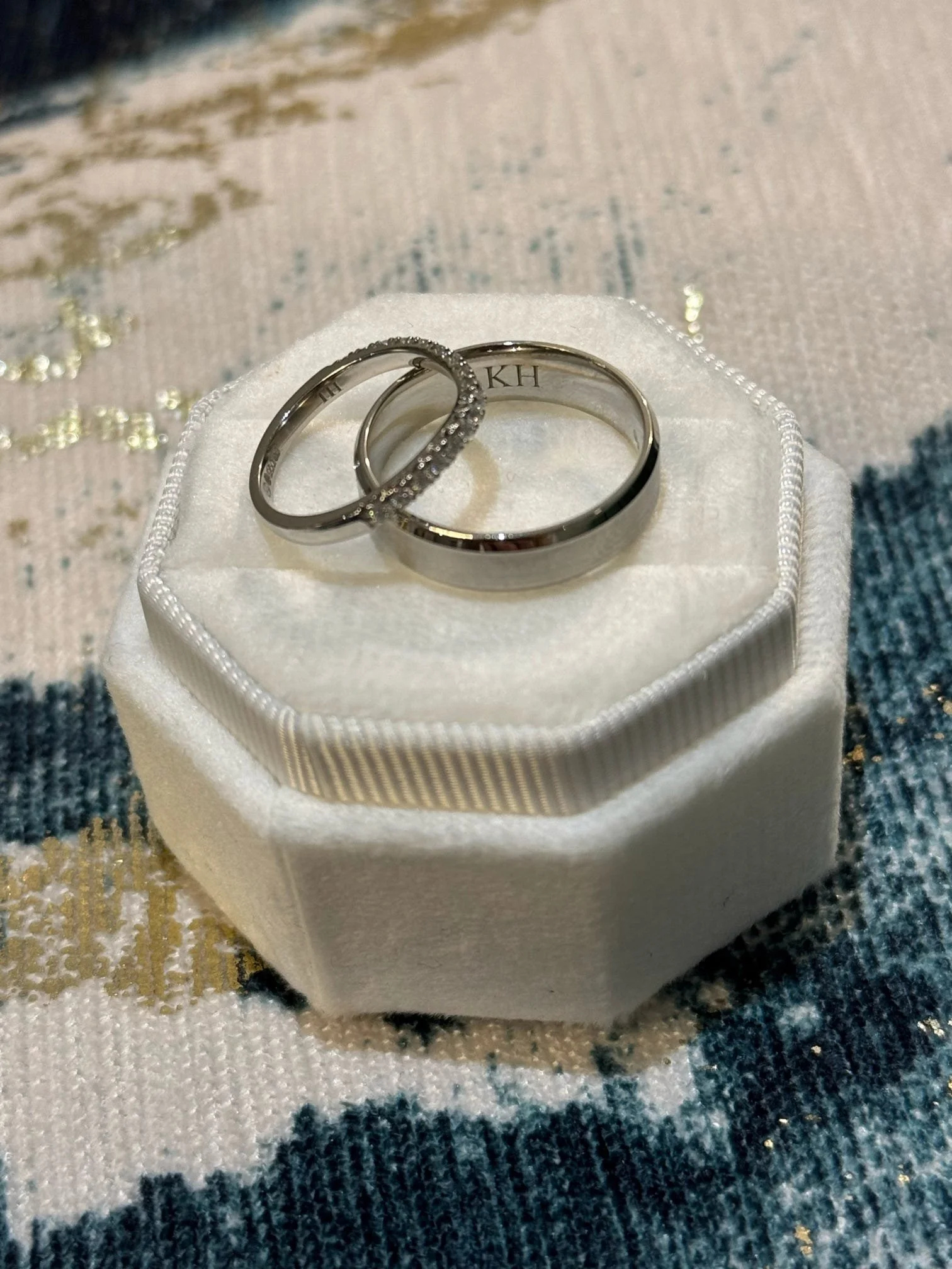 Two wedding rings, one with small diamonds, on a white ring pillow with a textured pattern. The pillow is on a fabric with a blue, black, white, and gold pattern.