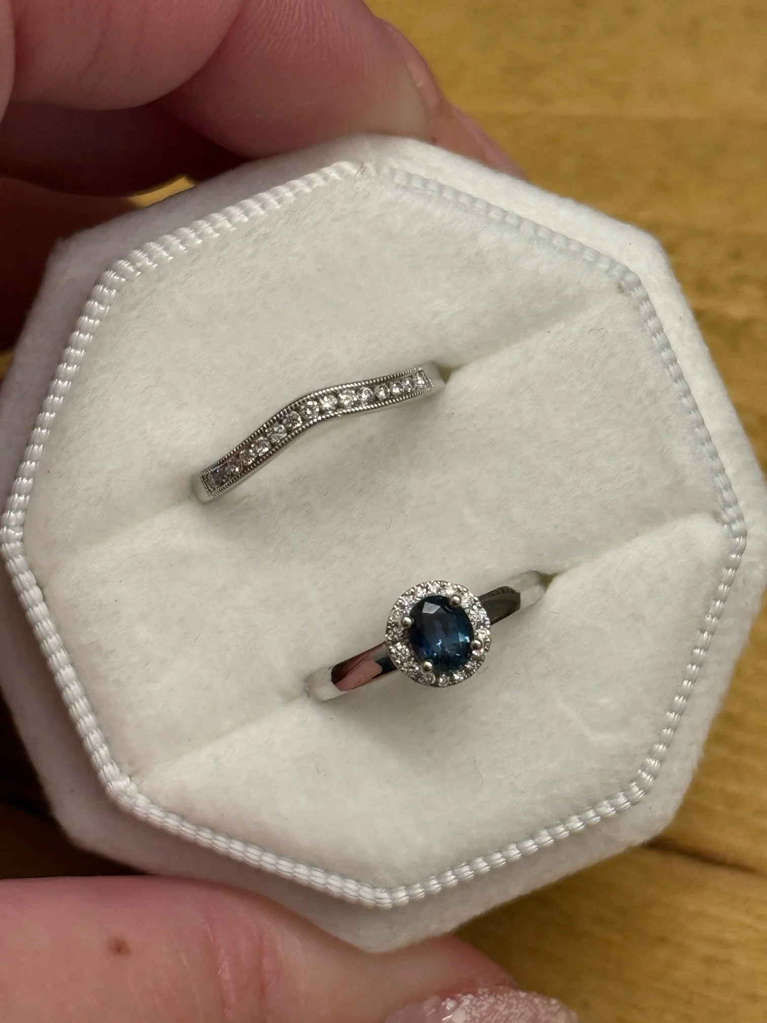 A white jewelry box containing a silver ring with a blue gemstone surrounded by small diamonds and an eternity band with small diamonds.