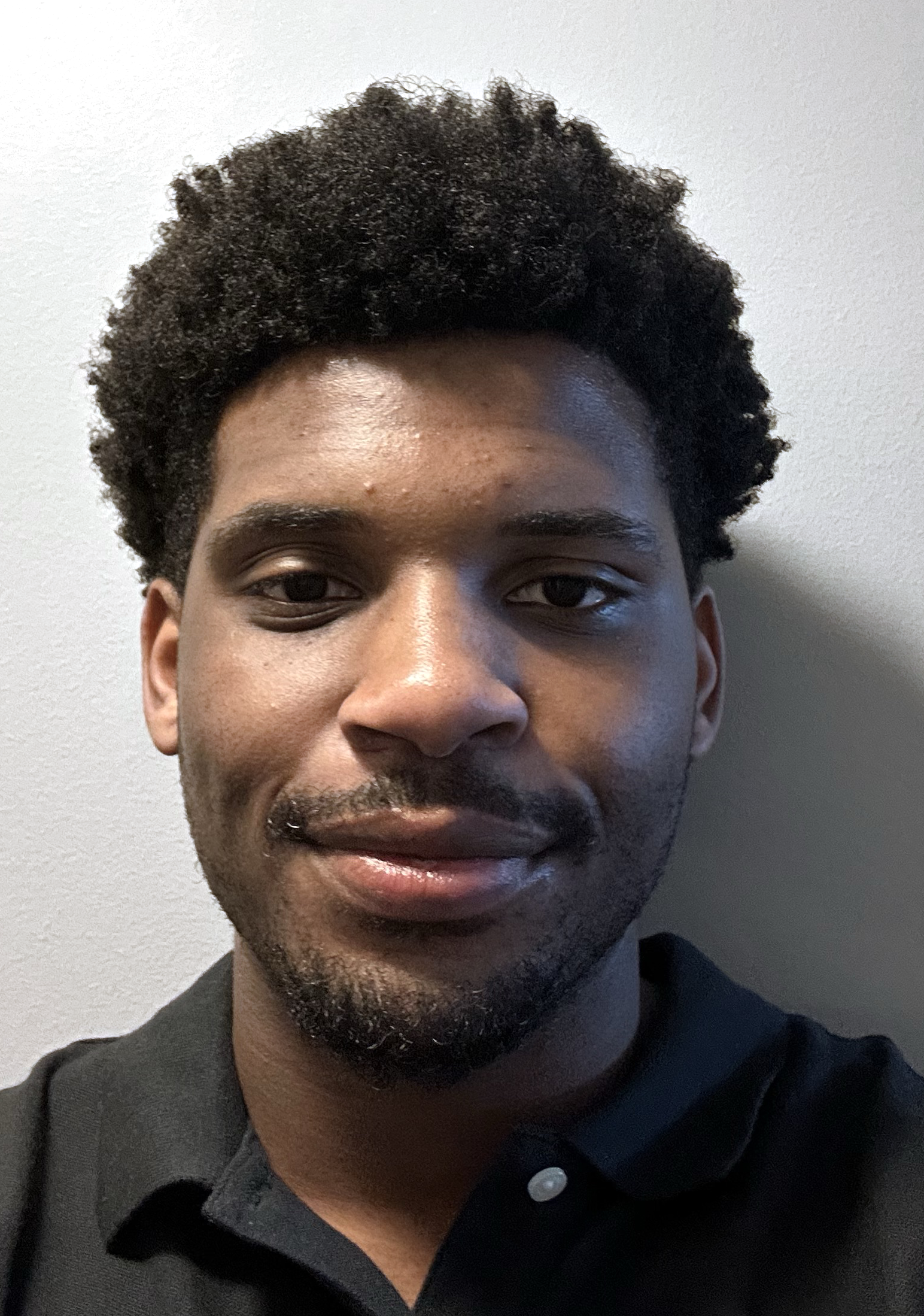 A close-up of a young man's face with black, textured hair, dark eyes, and a goatee, wearing a black collared shirt, against a background with digital glitch effects.