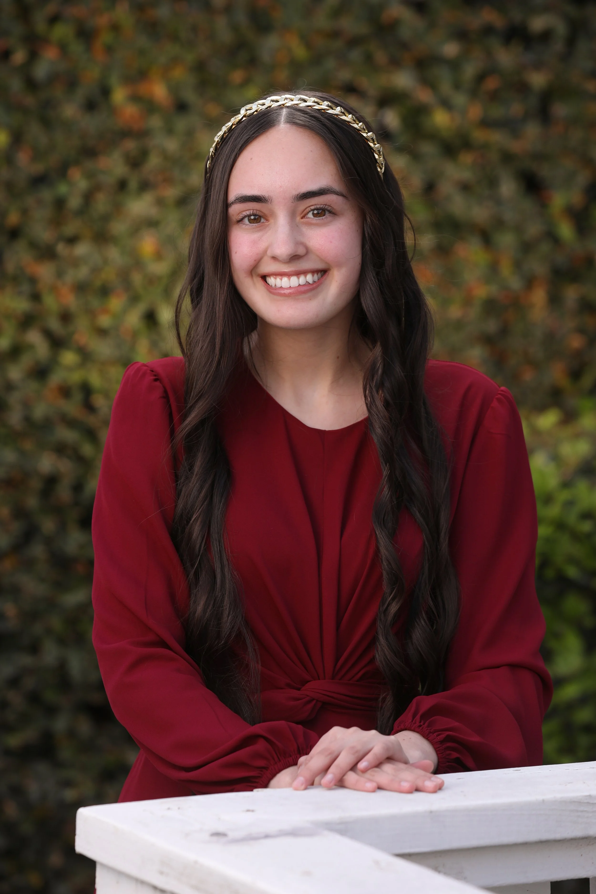 A young woman with long dark hair wearing a gold braided headband and a red long-sleeved dress, smiling, with her hands resting on a white wooden surface, outdoors with a blurred background of green foliage.