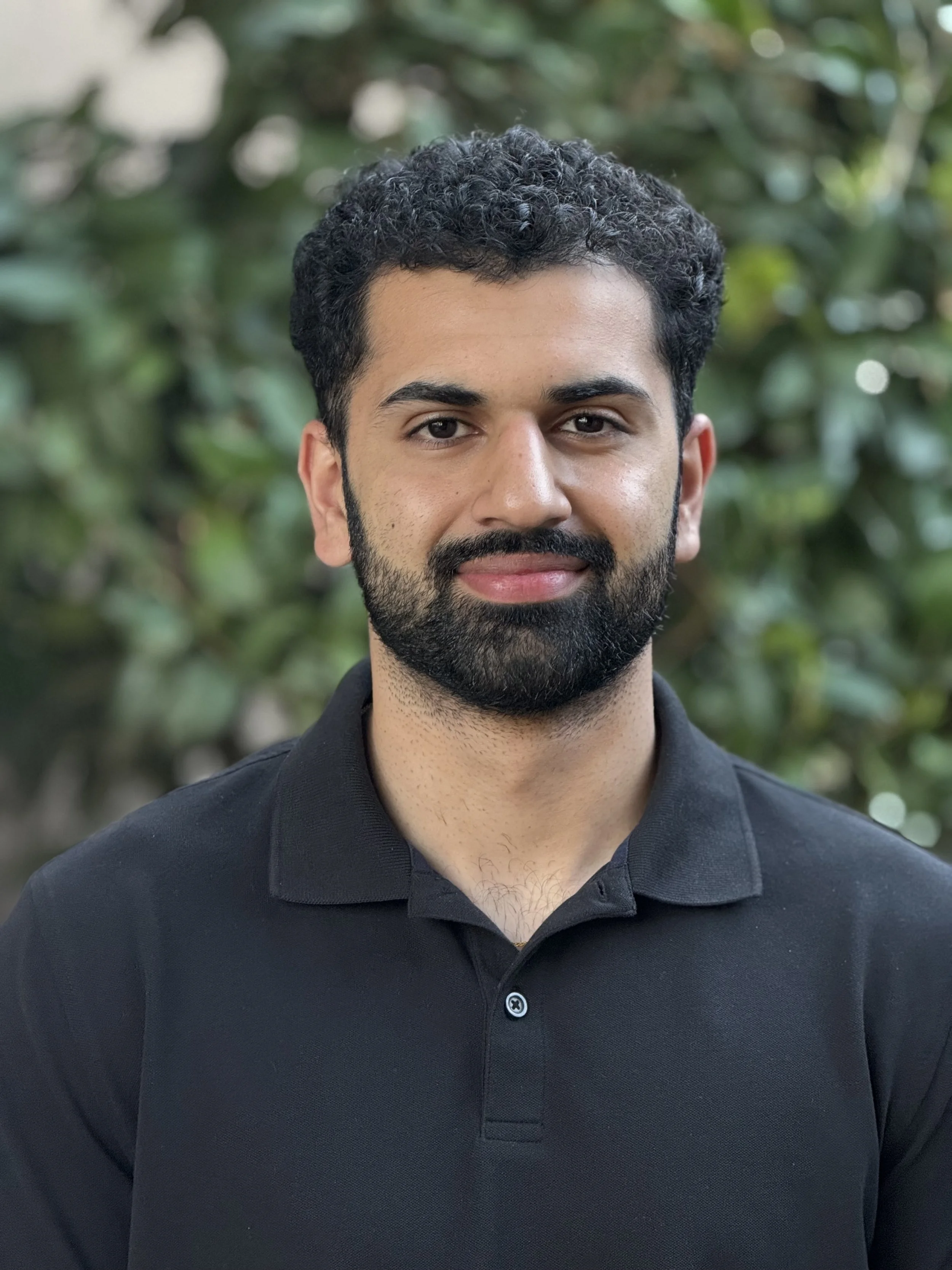 A man with curly dark hair, beard, and mustache wearing a black polo shirt, standing outdoors with greenery in the background.