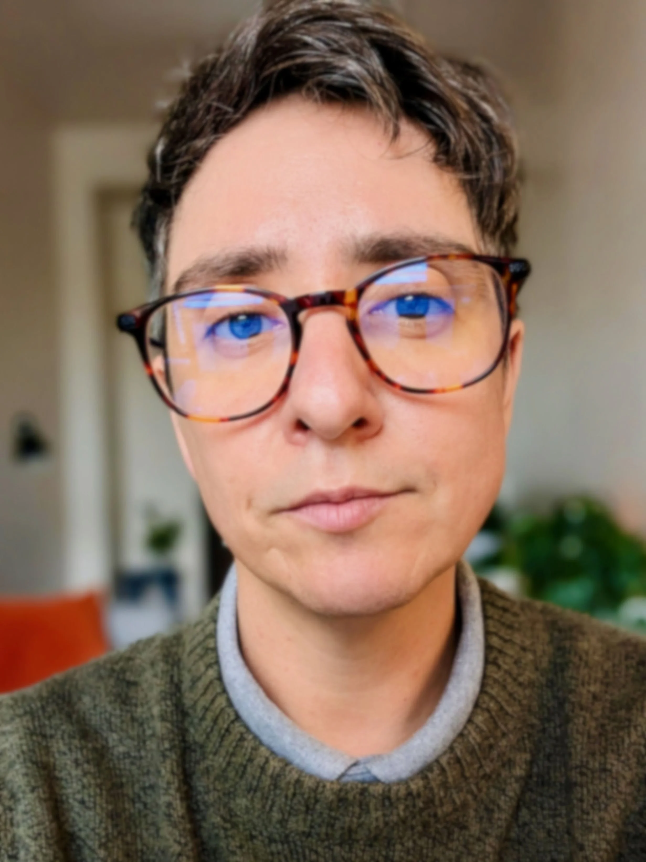 Close-up of a person with short dark hair and green eyes, wearing tortoiseshell glasses. They are dressed in an olive green sweater. The blurred background shows an office with several plants, a grey couch, and orange throw pillows.