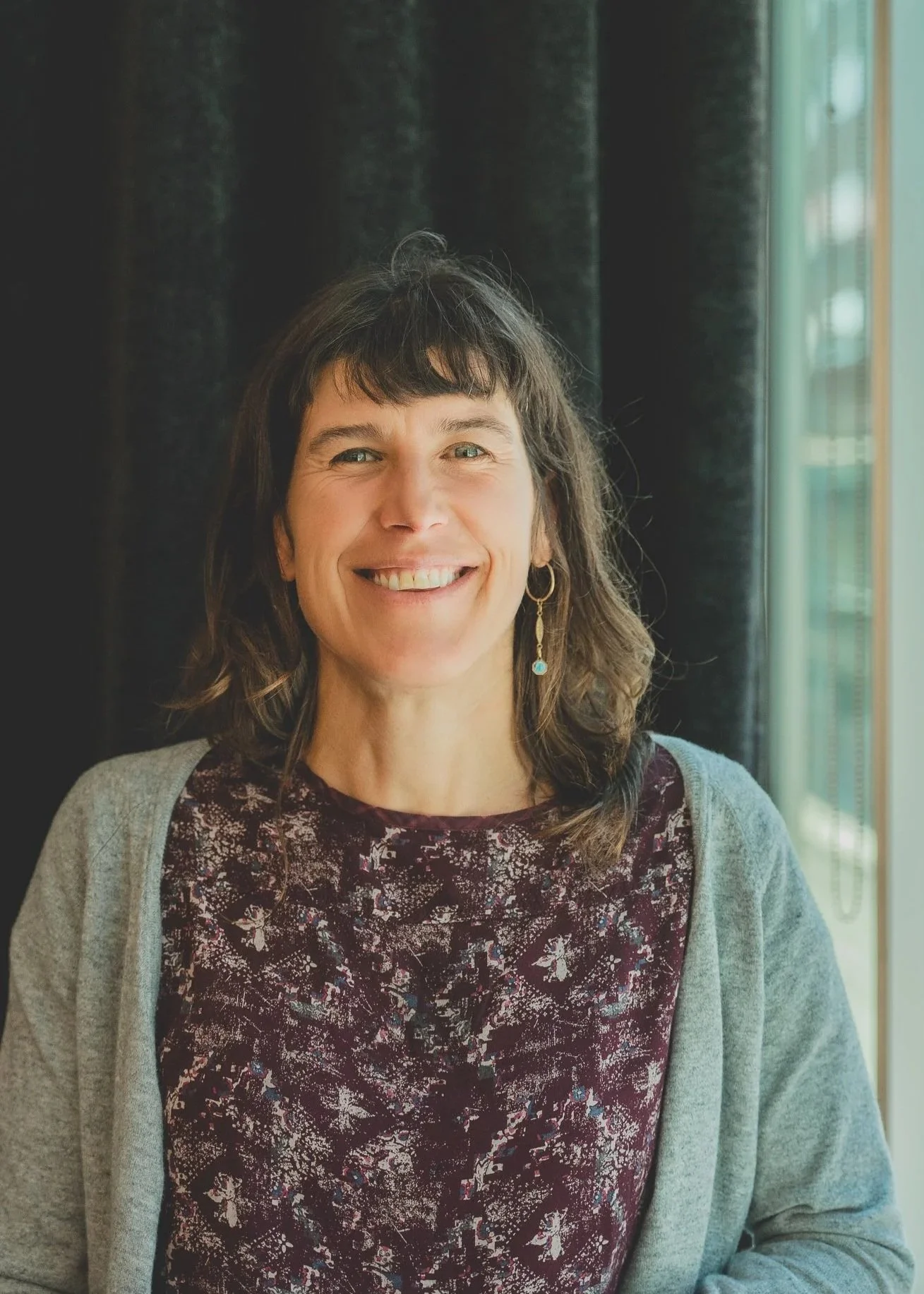 Femme souriante avec des cheveux bruns, portant une boucle d'oreille, un haut violet avec un motif floral et une veste grise, debout à côté d'une fenêtre avec rideaux foncés dans une pièce éclairée.