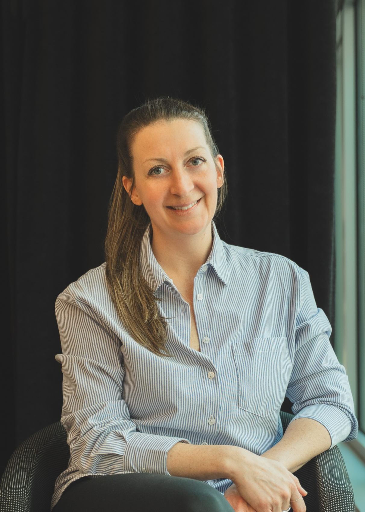 Une femme souriante assise près d'une fenêtre avec un fond noir.