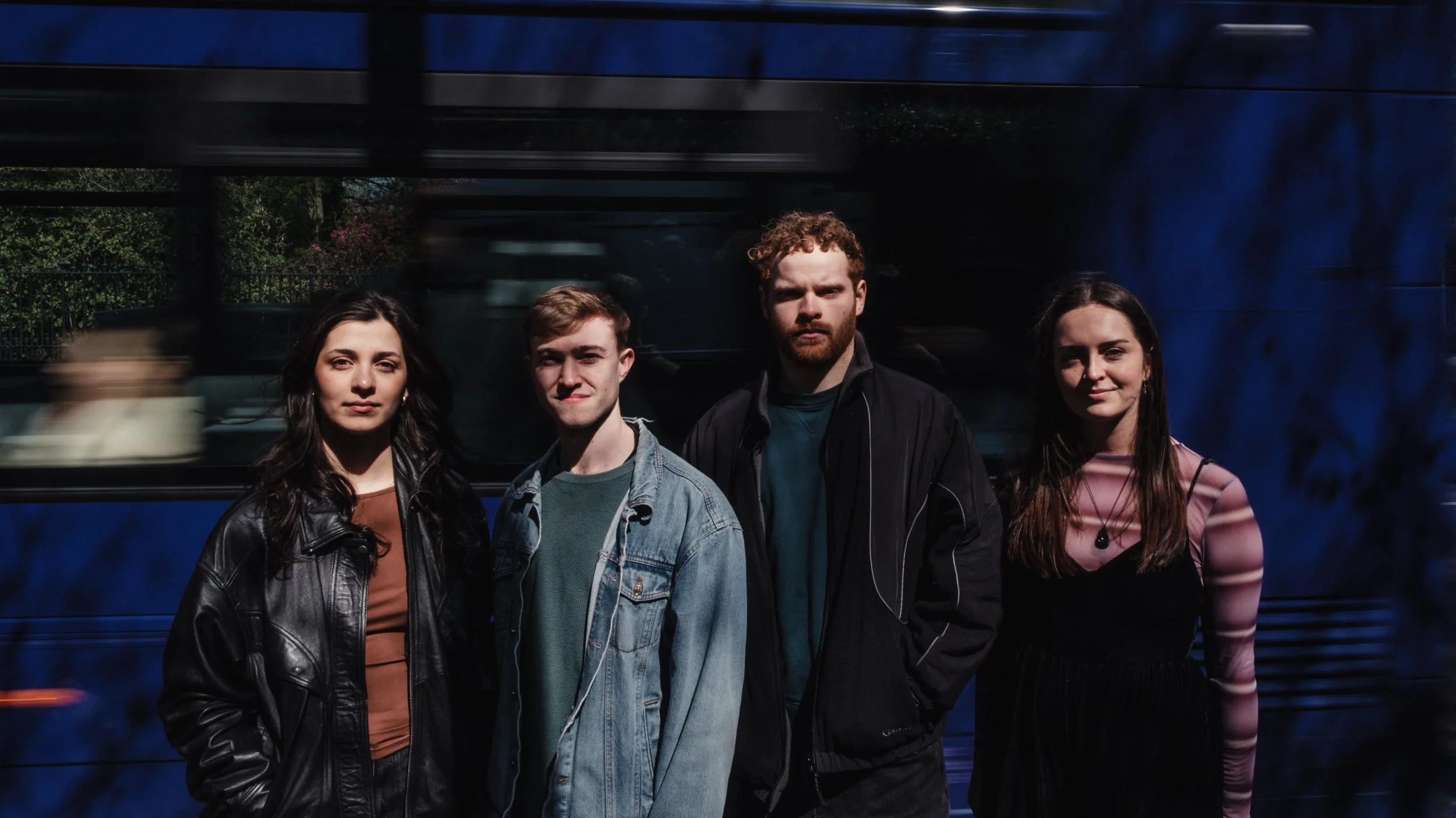 Four young adults standing in front of a blue bus, with blurred motion of the bus passing by behind them.