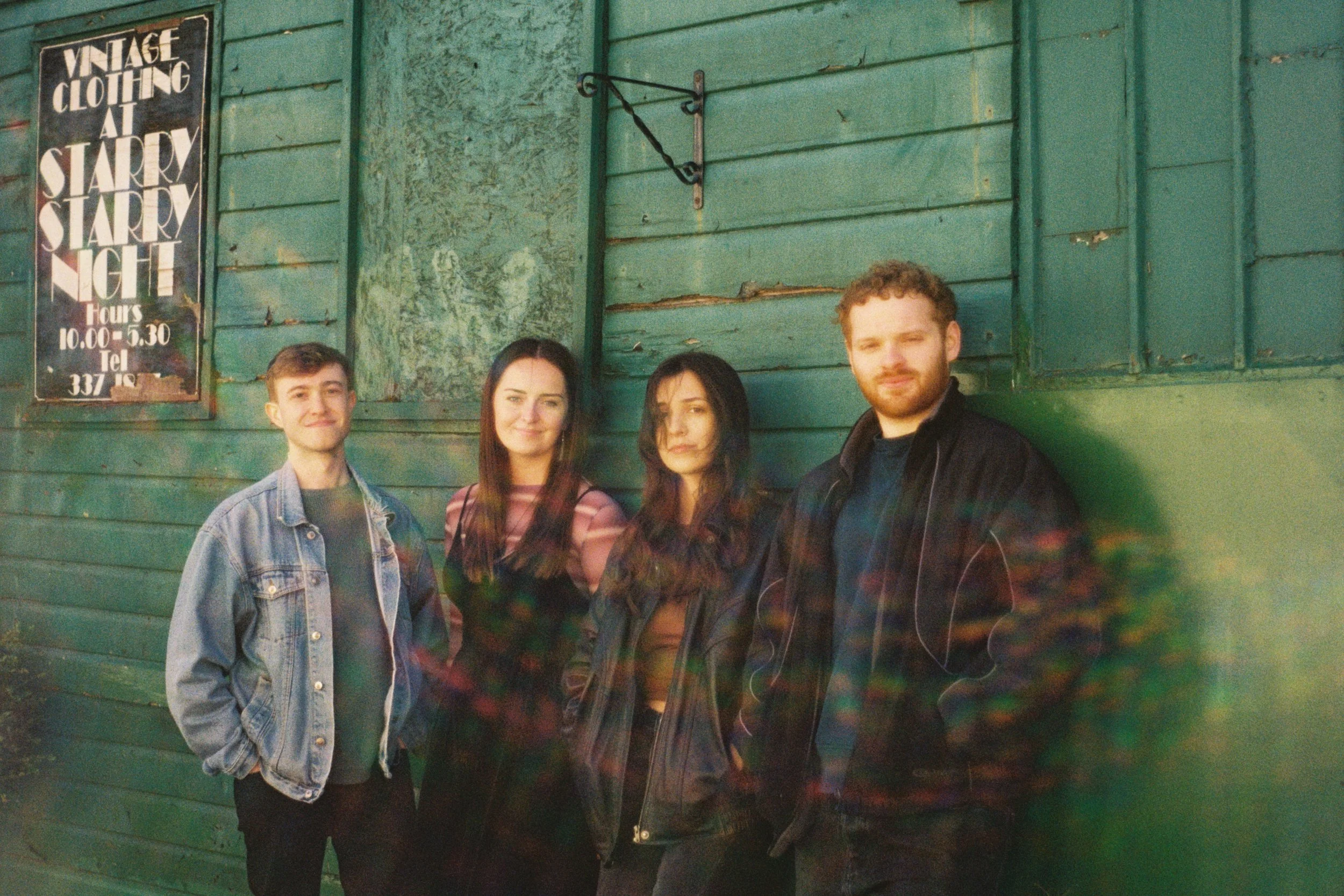 A group of four young adults standing in front of a weathered green wooden wall with a vintage clothing sign. The group includes two women and two men, all casually dressed, with relaxed expressions.
